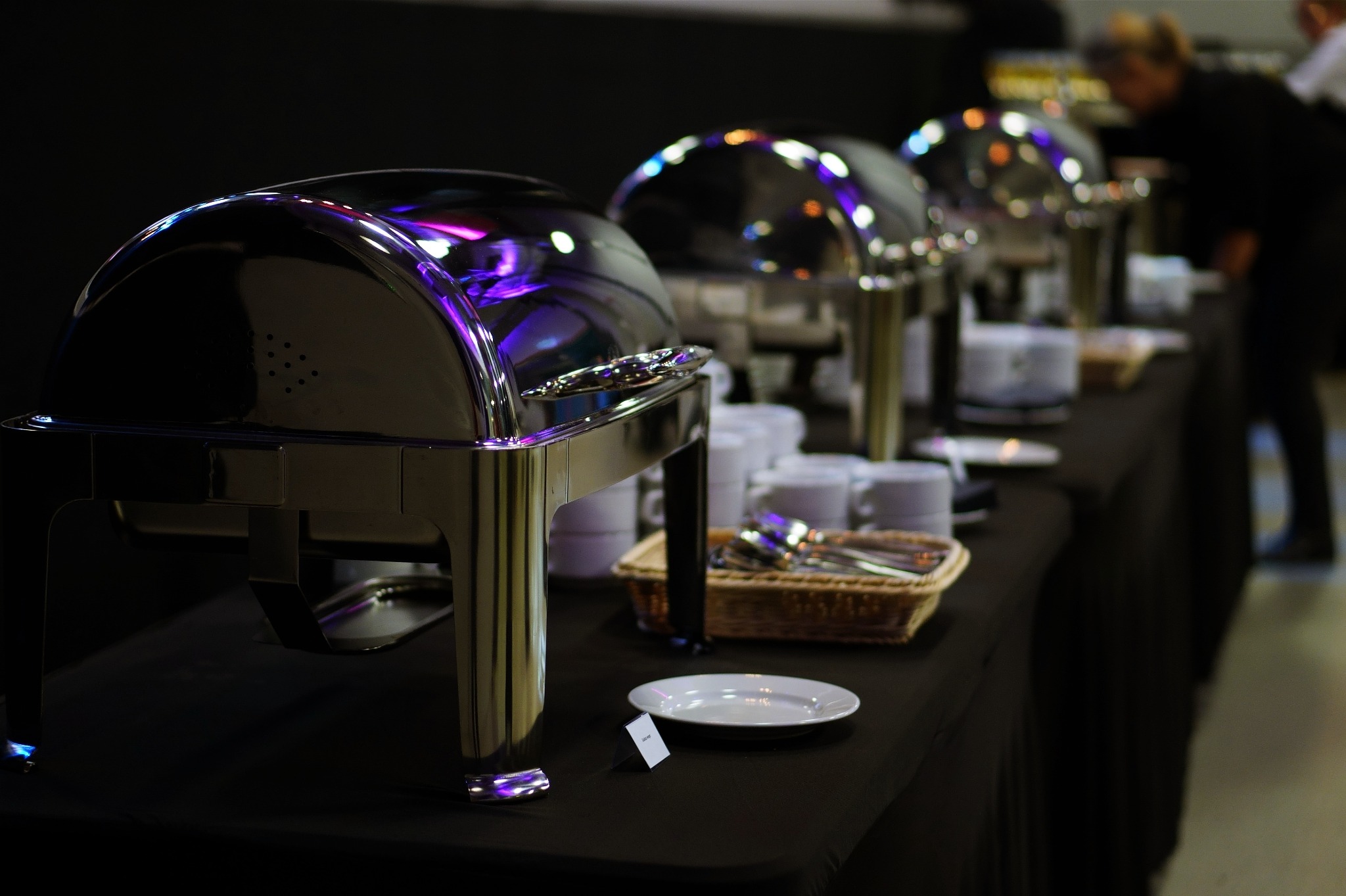 Stół cateringowy z podgrzewaczami bufetowymi, sztućcami w koszyku wiklinowym i stosem białych talerzy, oświetlony fioletowym światłem.