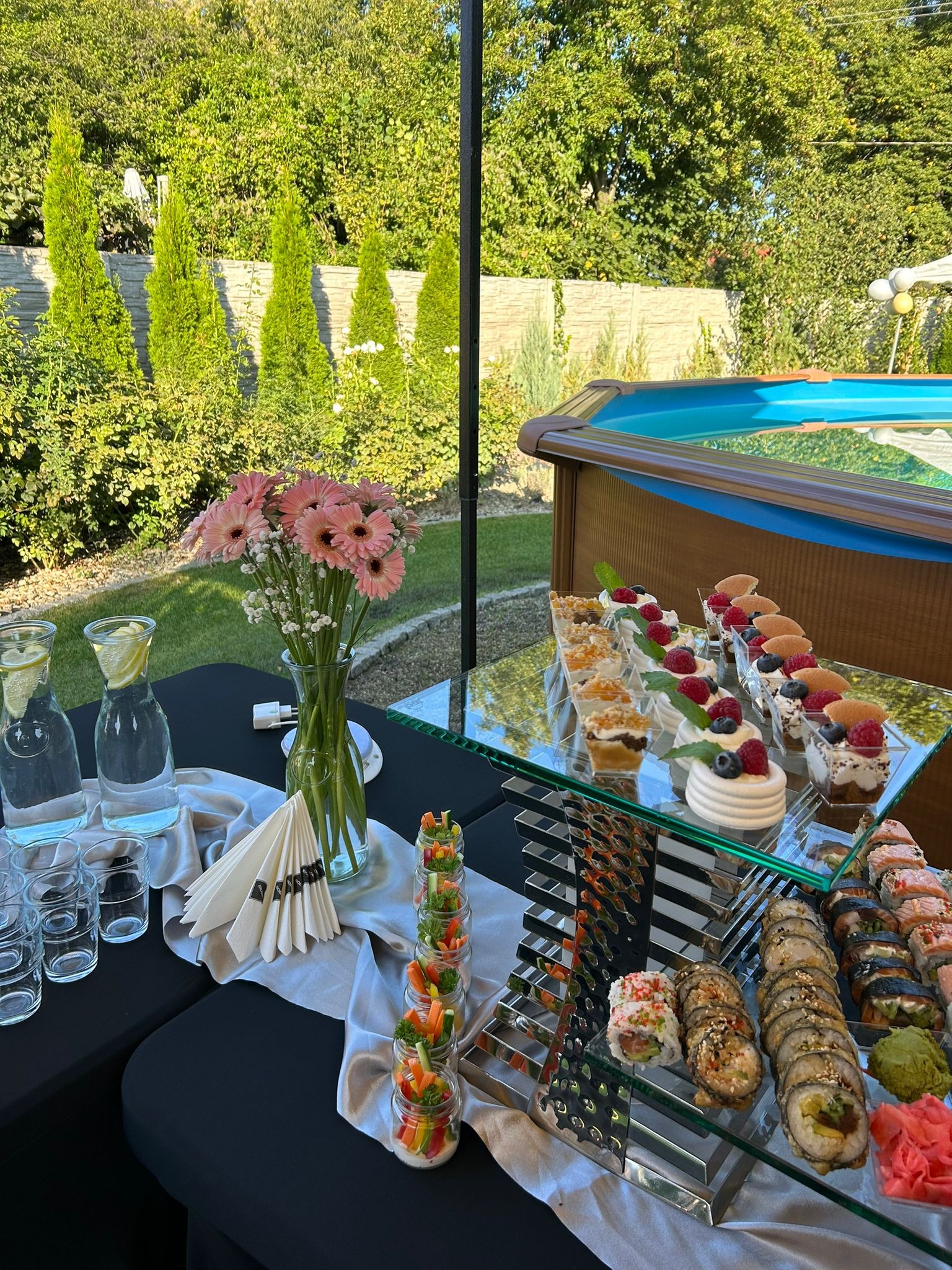 Stół cateringowy na zewnątrz z sushi, deserami i napojami w szklanych naczyniach, w tle ogród z basenem.