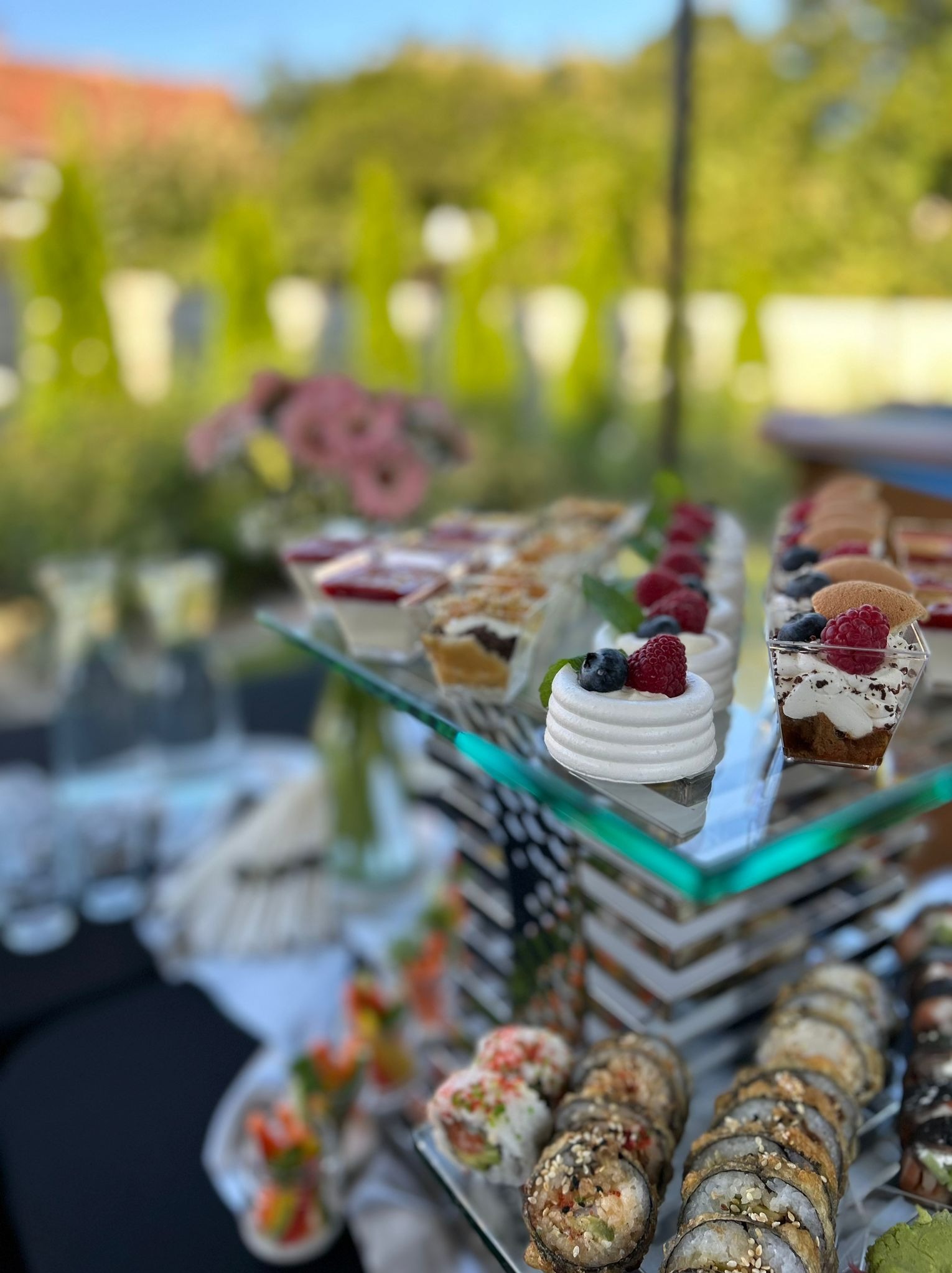 Szczegółowe ujęcie stołu z przekąskami na przyjęciu w plenerze: mini bezy z malinami i borówkami, deserki w przezroczystych pojemnikach, sushi i kwiaty w tle.
