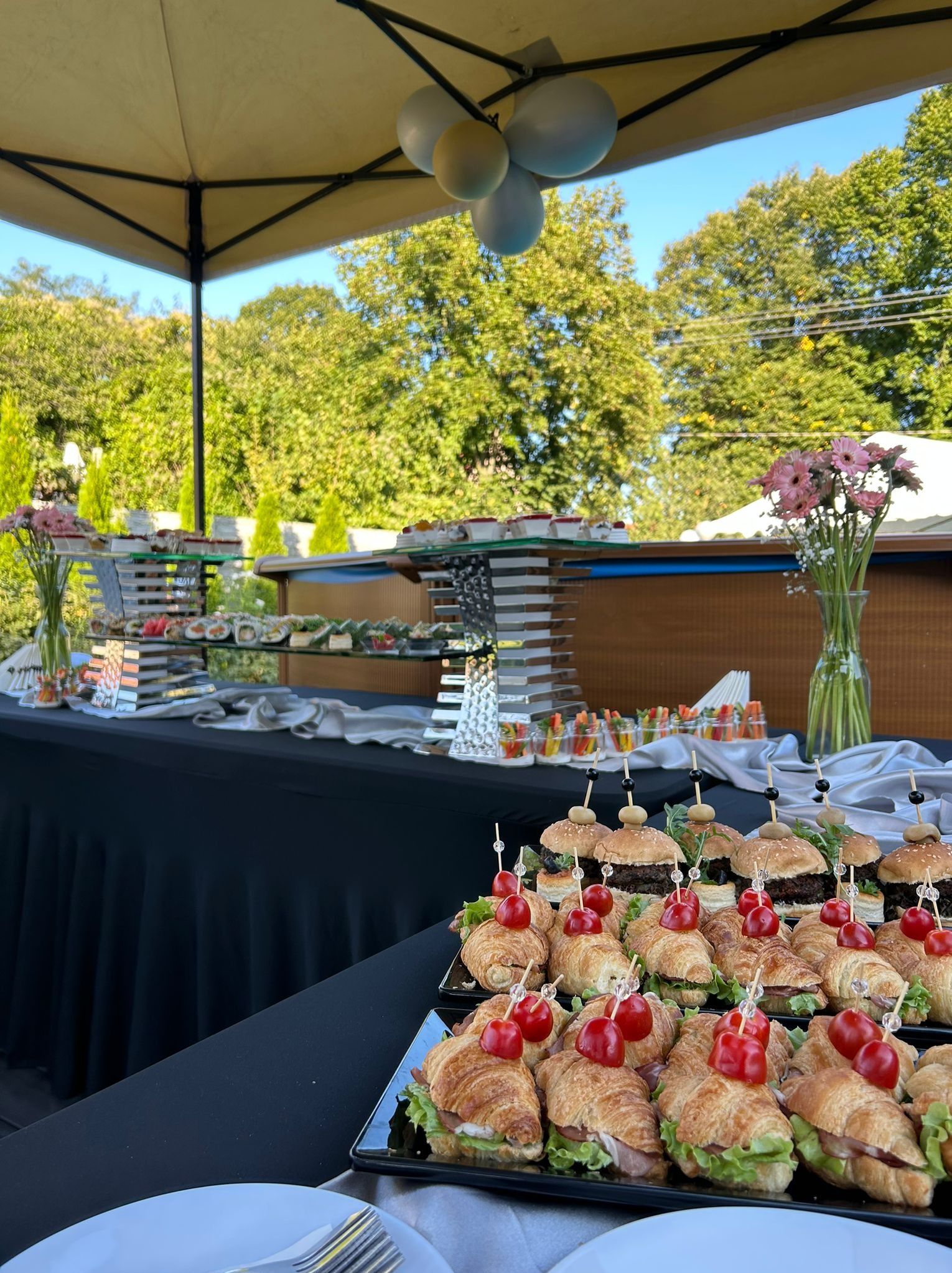 Stół cateringowy na zewnątrz z przekąskami: mini croissanty z szynką i sałatą, mini burgery z oliwkami, sushi, warzywne przekąski w słoiczkach, dekoracja kwiatowa i balony.