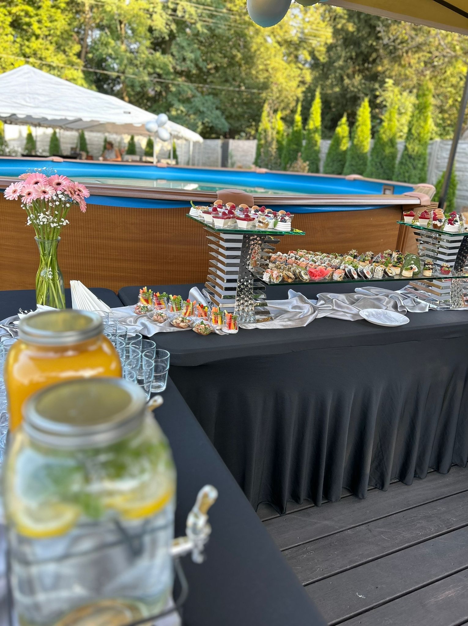 Stół z czarnym obrusem, na którym znajdują się przekąski, sushi, desery, napoje w słojach z kranikami, szklanki, wazon z różowymi kwiatami, w tle basen i zielone drzewa.