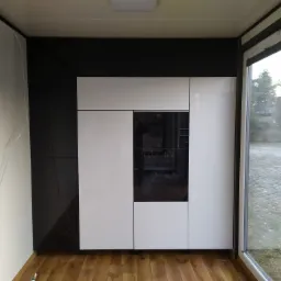 Nowoczesna zabudowa kuchenna z białymi i czarnymi frontami, wbudowany piekarnik, widok na ogród przez okno.