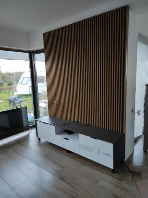 Nowoczesna szafka RTV w kolorze białym i czarnym z dekoracyjnym panelem ściennym z pionowych, drewnianych listewek, na tle okna z widokiem na ogród.