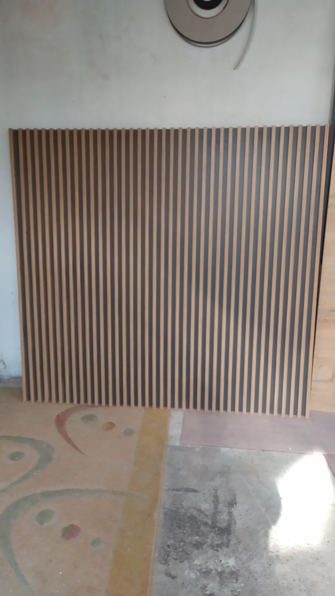 Panel ścienny z pionowych listew w kolorze drewna i grafitu, ustawiony na podłodze z wzorami, widoczne niedoskonałości podłoża.