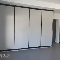 Cztery przesuwne drzwi szafy wnękowej z szarymi panelami i czarnymi ramami, widoczne z niskiego kąta, z fragmentem drugiej szafy po prawej i szarą podłogą.