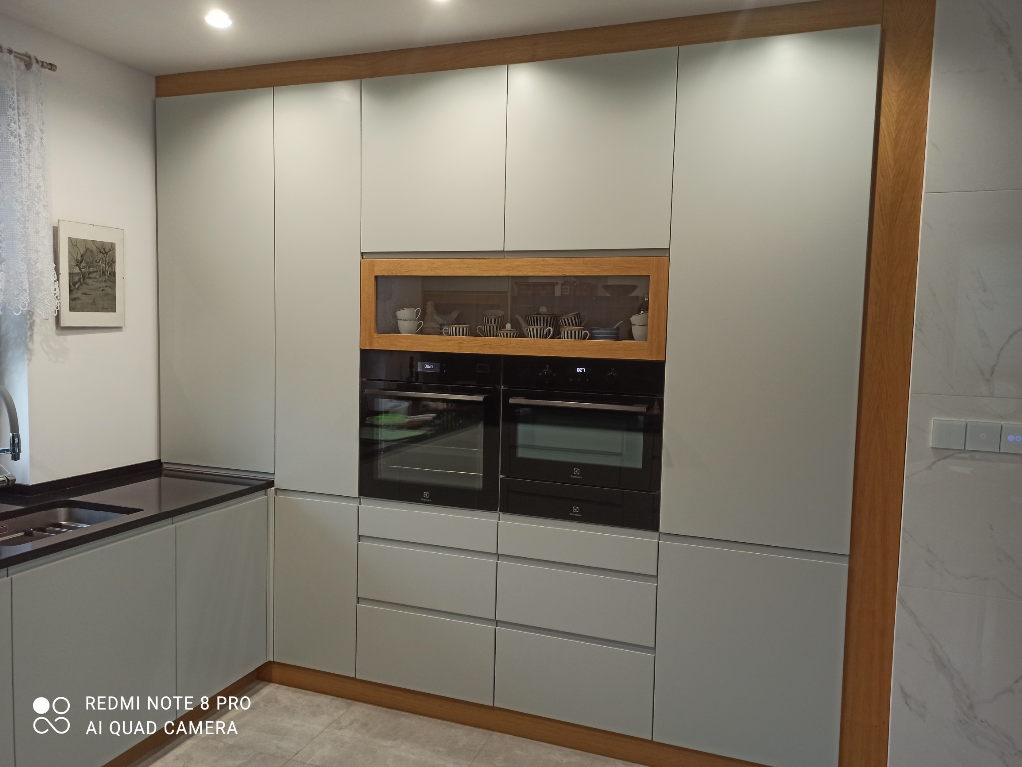 Szare szafki kuchenne z drewnianymi akcentami, dwie wbudowane czarne piekarniki i przeszklona szafka z filiżankami i czajnikiem.