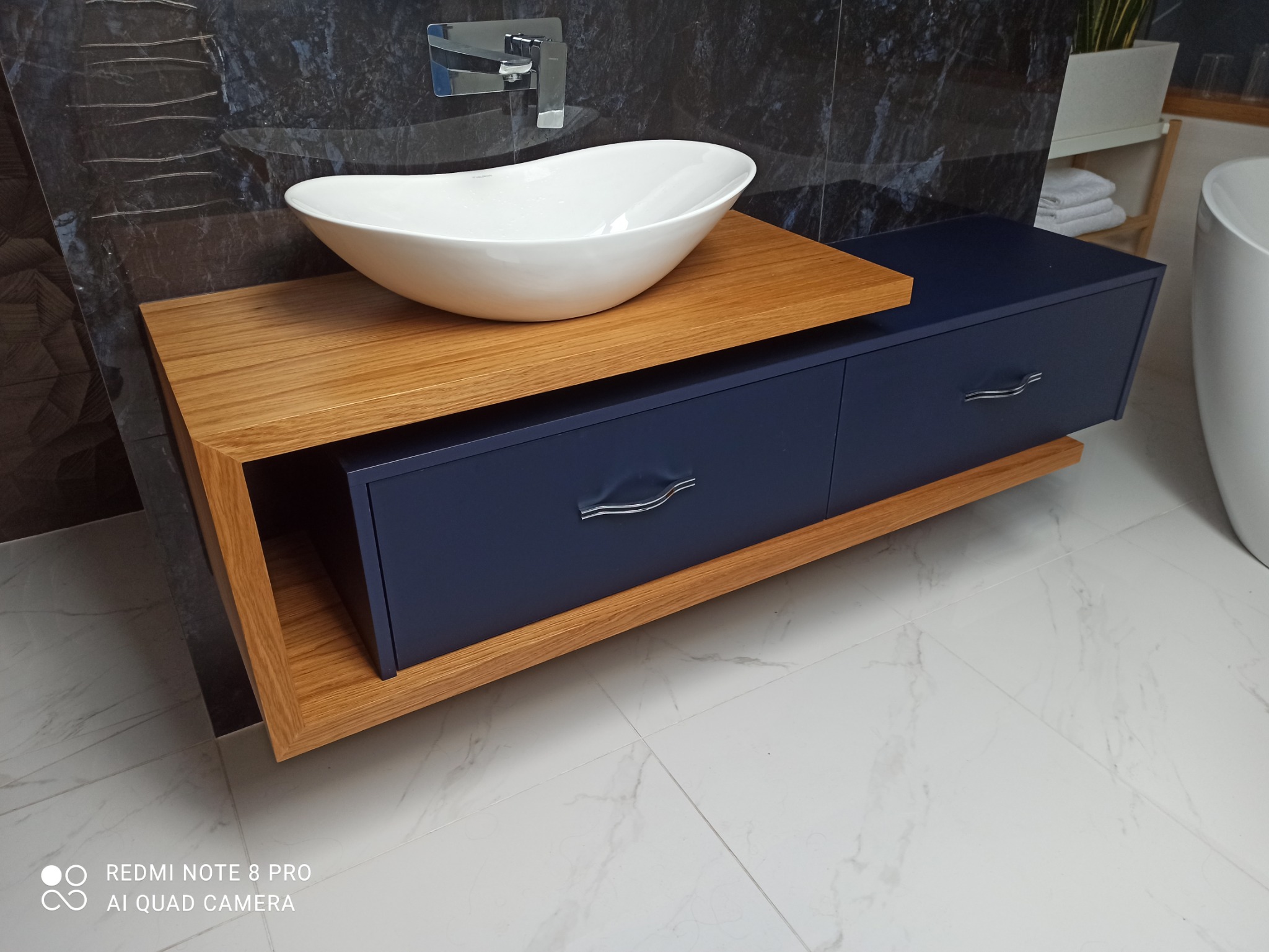 Nowoczesna łazienka z umywalką nablatową w kształcie łzy na drewnianym blacie z szafką w kolorze granatowym, marmurowe płytki na ścianie i podłodze, minimalistyczna bateria umywalkowa.