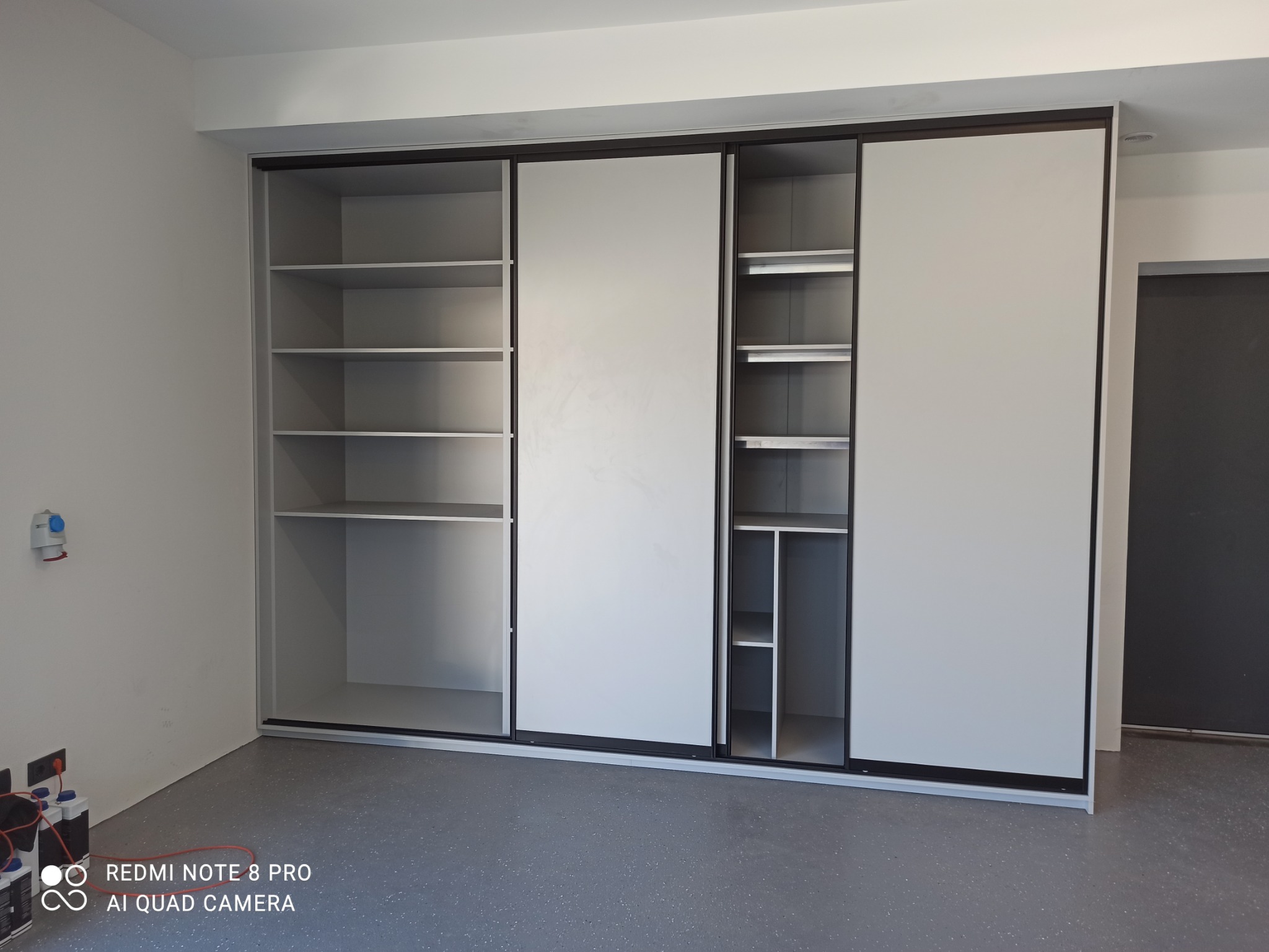 Szafa wnękowa z przesuwnymi drzwiami w kolorze jasnoszarym, z widocznymi półkami w otwartej części, na tle białej ściany z instalacją elektryczną i szarej podłogi.