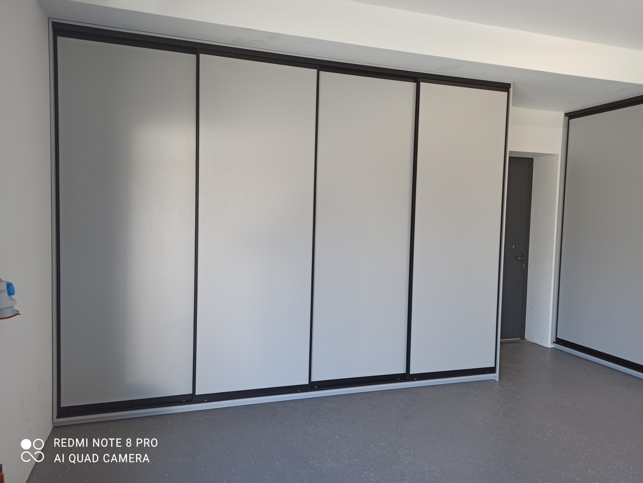 Cztery przesuwne drzwi szafy wnękowej z szarymi panelami i czarnymi ramami, widoczne z niskiego kąta, z fragmentem drugiej szafy po prawej i szarą podłogą.