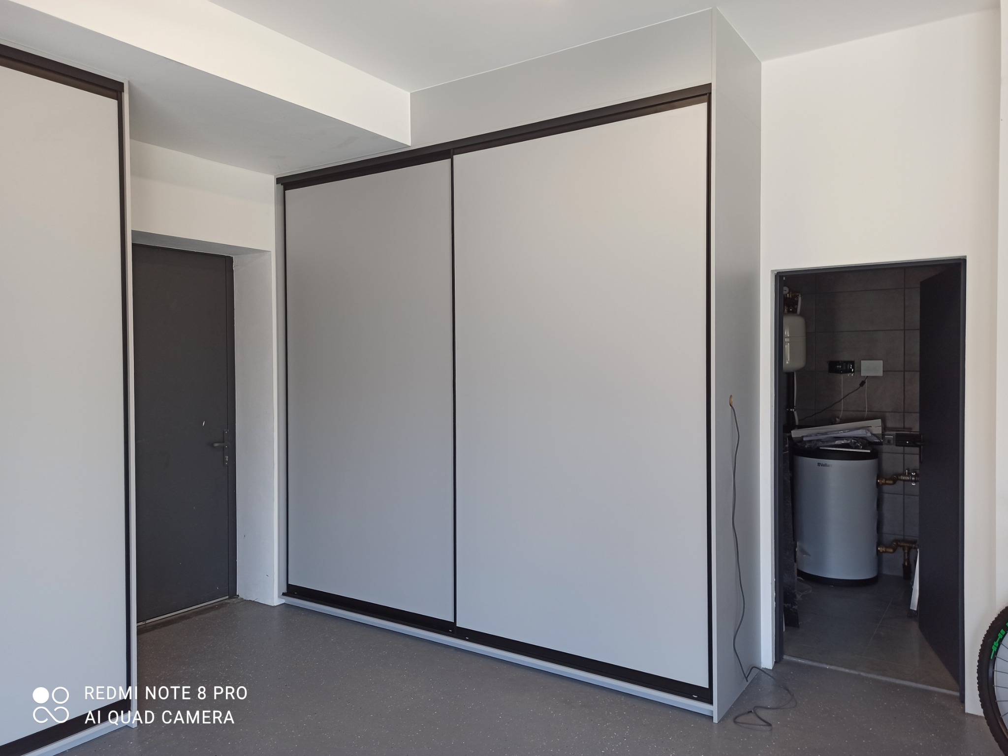 Szara szafa przesuwna z czarnymi obramowaniami, w tle widoczne drzwi i pomieszczenie z bojlerem, minimalistyczny design.