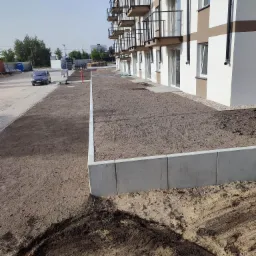 Przygotowany teren pod trawnik wzdłuż nowego budynku wielorodzinnego z betonowymi obrzeżami i podsypką z piasku.