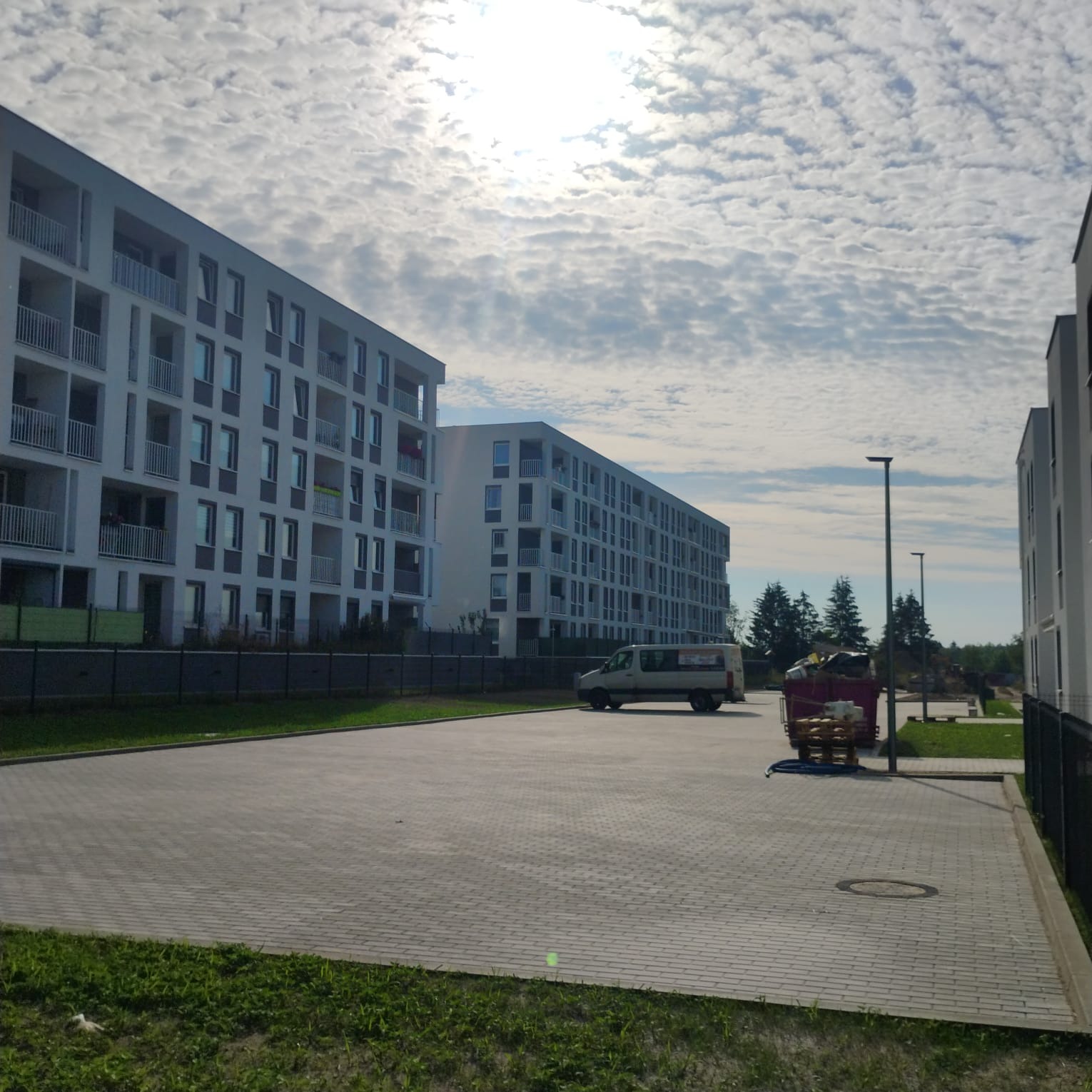 Nowe osiedle w Skórzcu z wybrukowanym parkingiem, widoczny kontener na śmieci i zaparkowany bus.