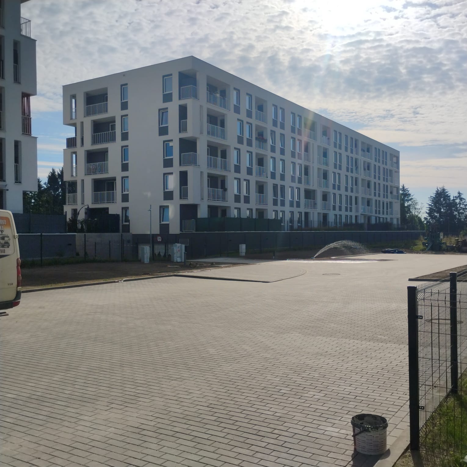 Nowo wybrukowany plac przed nowoczesnym budynkiem mieszkalnym, zraszacz nawadnia trawnik, widoczny częściowo zaparkowany samochód dostawczy.