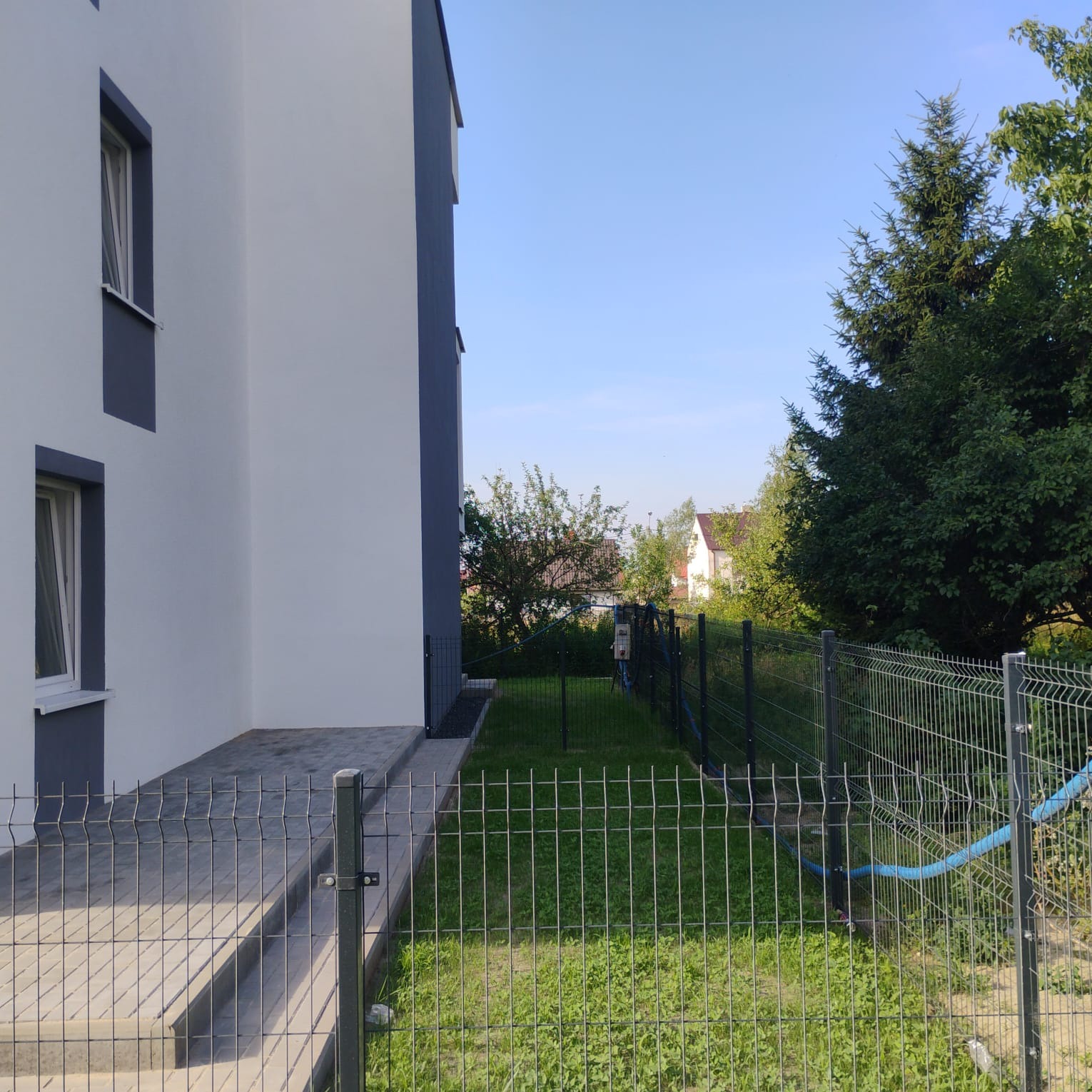 Nowoczesny dom z szarą elewacją i ułożoną kostką brukową na podejściu, widoczna metalowa siatka ogrodzeniowa i trawnik.