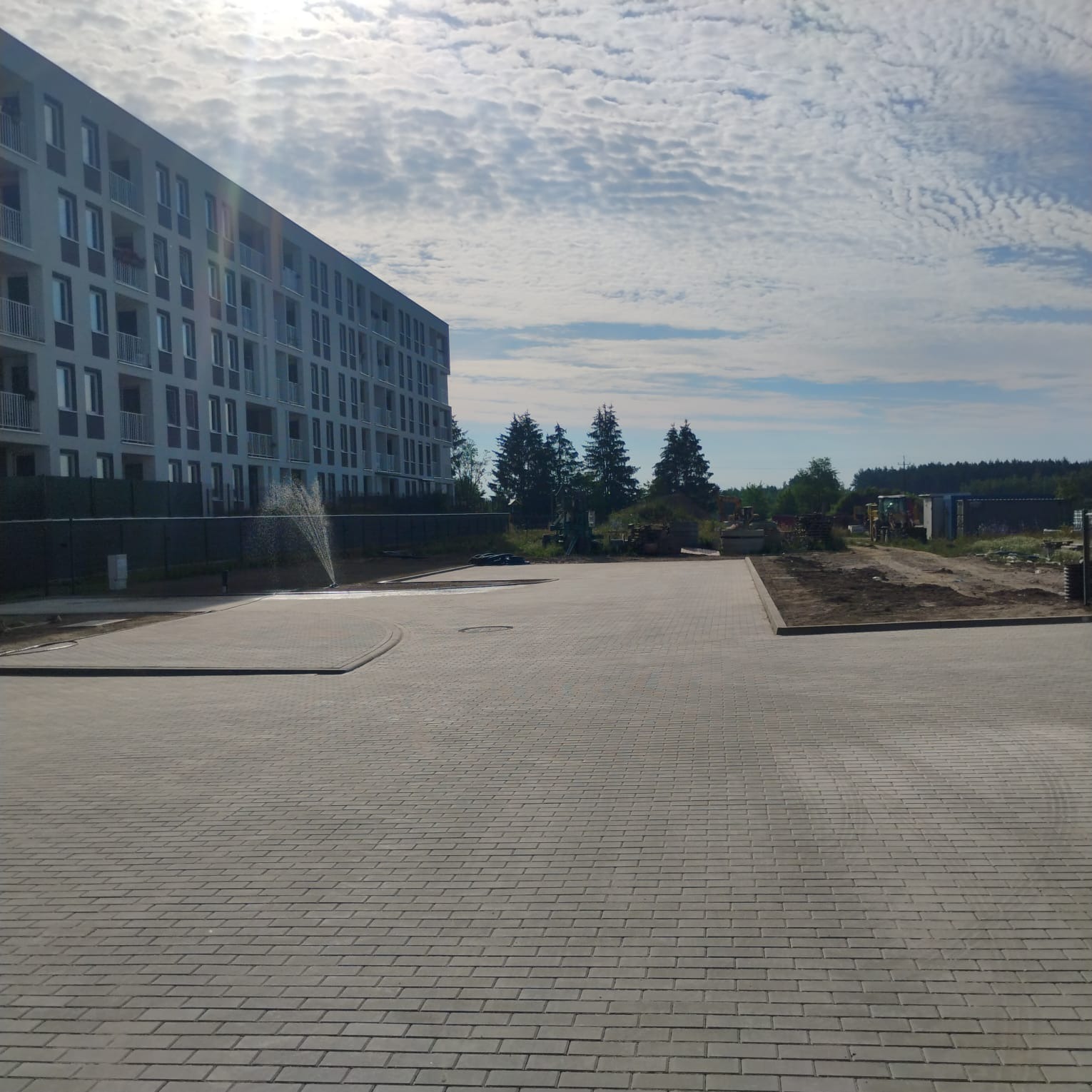 Rozległy plac wyłożony szarą kostką brukową przed nowoczesnym budynkiem mieszkalnym, w tle widoczne drzewa i fragment terenu przygotowanego pod dalsze prace, zraszacz nawadnia część placu.