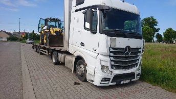 Ciężarówka Mercedes-Benz Actros z naczepą niskopodwoziową przewożąca żółtą koparko-ładowarkę, widok z boku na tle zieleni i zabudowań.