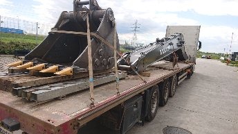 Transport ciężarowy: Łyżka koparki z zębami i ramię koparki zabezpieczone pasami na niskopodwoziowej naczepie ciężarowej, widoczne elementy konstrukcyjne i słupki ogrodzenia w tle.