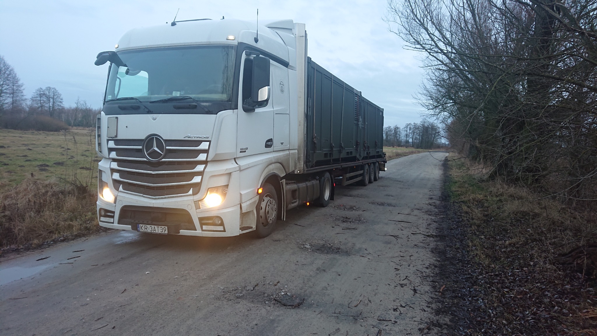 Biały ciągnik siodłowy Mercedes-Benz Actros z naczepą kontenerową na polnej drodze w pochmurny dzień, widok z przodu, tablica rejestracyjna widoczna.