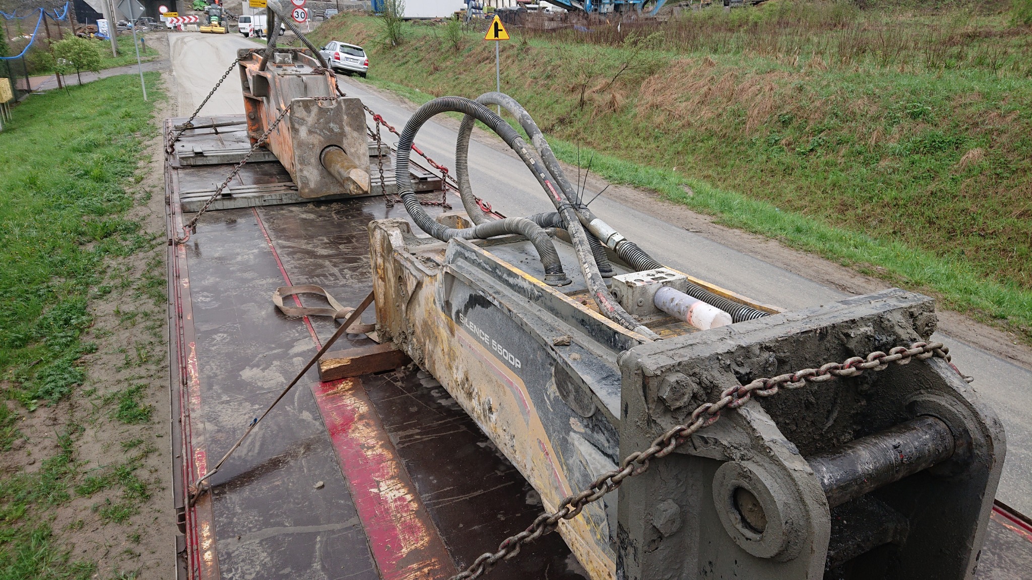 Hydrauliczny młot wyburzeniowy Silence 5500P, zabezpieczony łańcuchami, transportowany na niskopodwoziowej platformie ciężarowej, widok z góry na tle wiejskiego krajobrazu z drogą i znakami drogowymi.