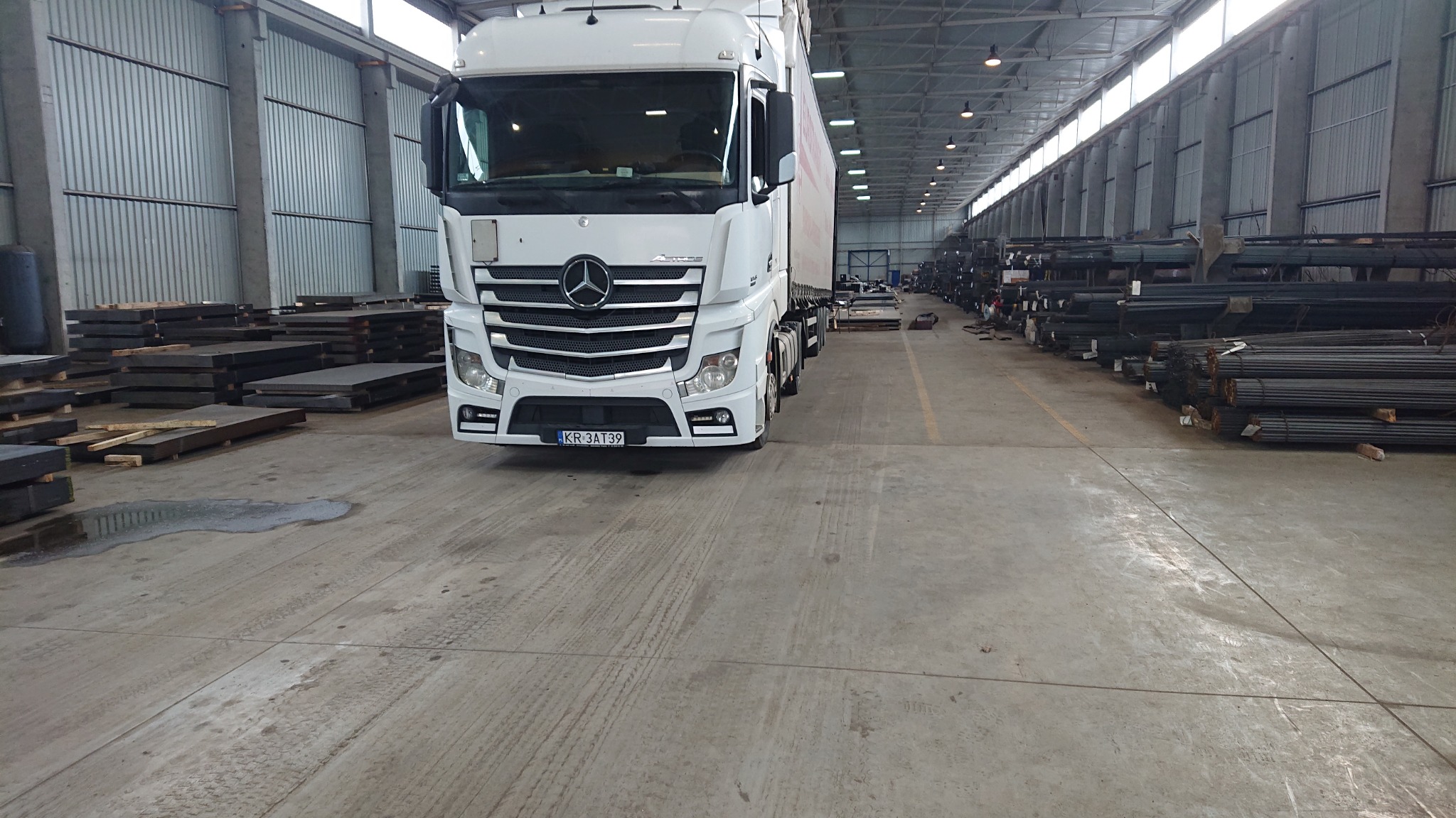 Biała ciężarówka Mercedes Actros z widoczną tablicą rejestracyjną KR 3AT39 stoi w hali magazynowej wypełnionej materiałami budowlanymi, takimi jak stalowe pręty i płyty.