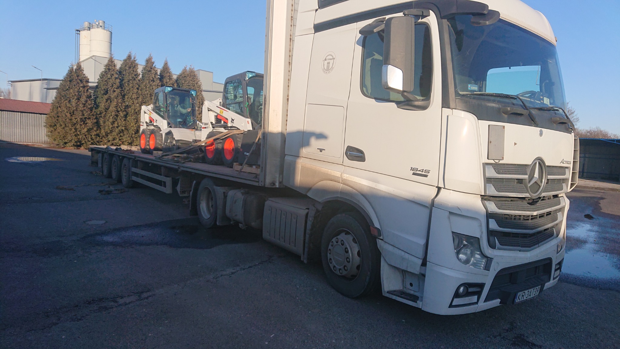 Ciężarówka Mercedes Actros 1845 przewozi dwie białe miniładowarki Bobcat z pomarańczowymi kołami na niskopodwoziowej platformie transportowej.