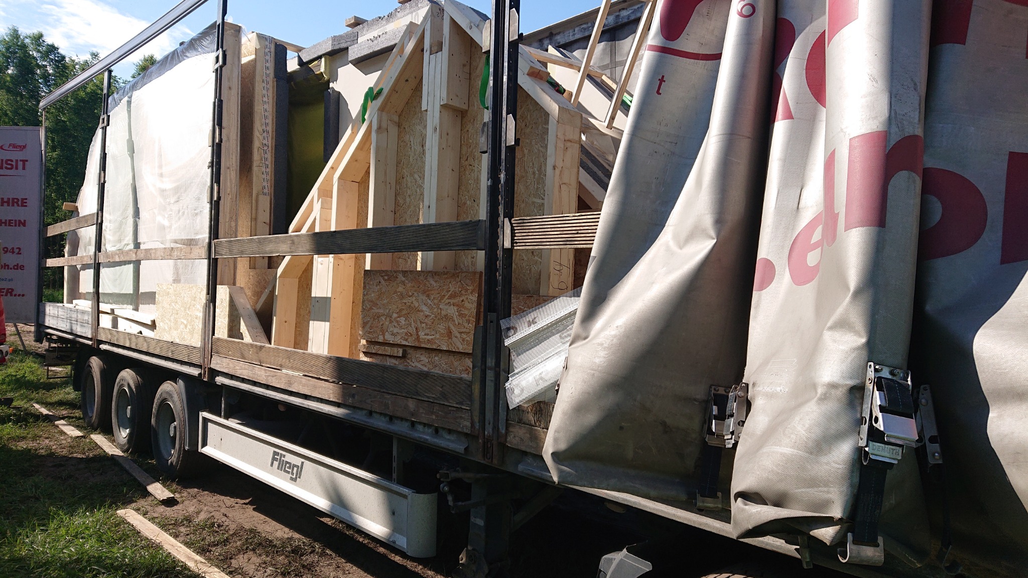 Ciężarówka marki Fliegl załadowana elementami konstrukcyjnymi drewnianych domów modułowych, zabezpieczona plandeką z różowymi napisami, widok z boku na trawiastym podłożu.