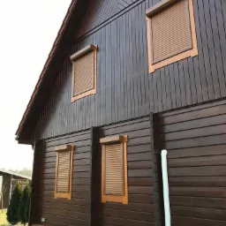 Brązowy drewniany dom z zamontowanymi zewnętrznymi roletami w kolorze zbliżonym do koloru okien i elewacji, z widocznymi klimatyzatorami na zewnątrz.