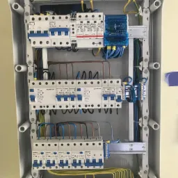 Otwarta skrzynka elektryczna z bezpiecznikami, listwami zaciskowymi i przewodami o różnych kolorach, zamontowana wewnątrz jasnej obudowy.