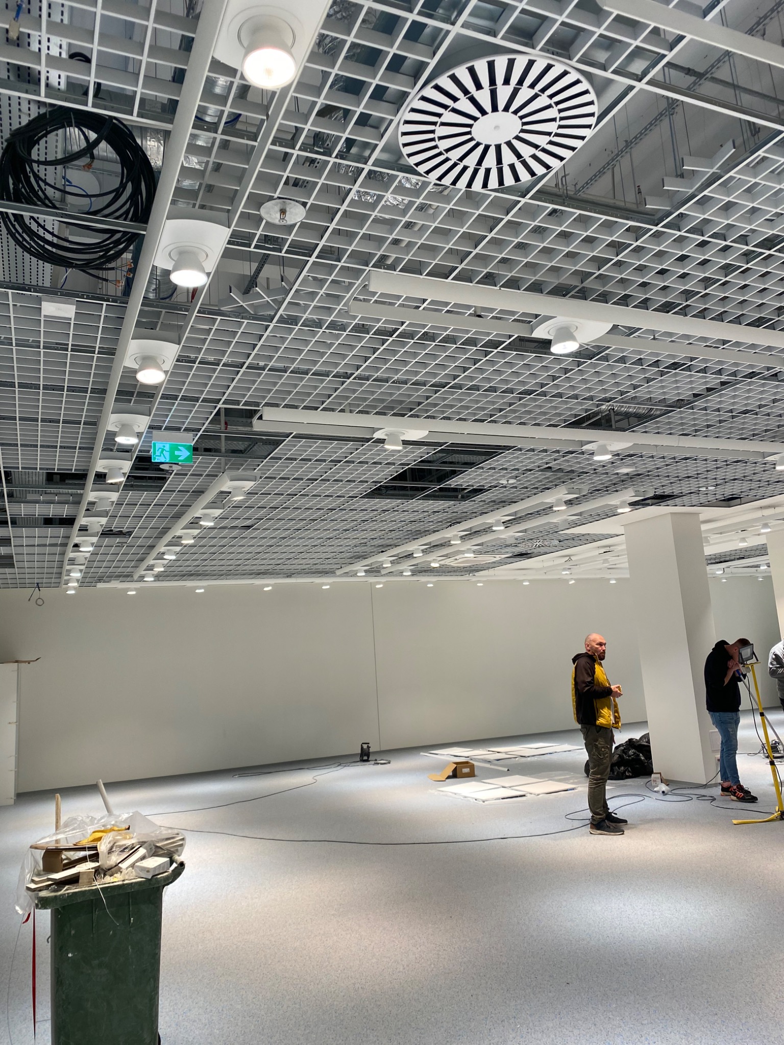 Montaż sufitu podwieszanego z instalacją oświetleniową i wentylacyjną w przestrzeni biurowej, widoczni pracownicy i śmietnik z odpadami.