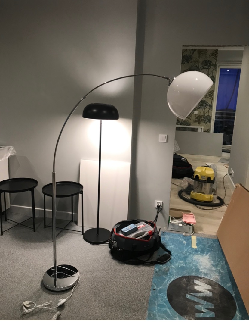 Dwie lampy stojące, czarna i srebrna z białym kloszem, w pokoju podczas remontu; widoczny otwarty worek z narzędziami i odkurzacz przemysłowy w tle.