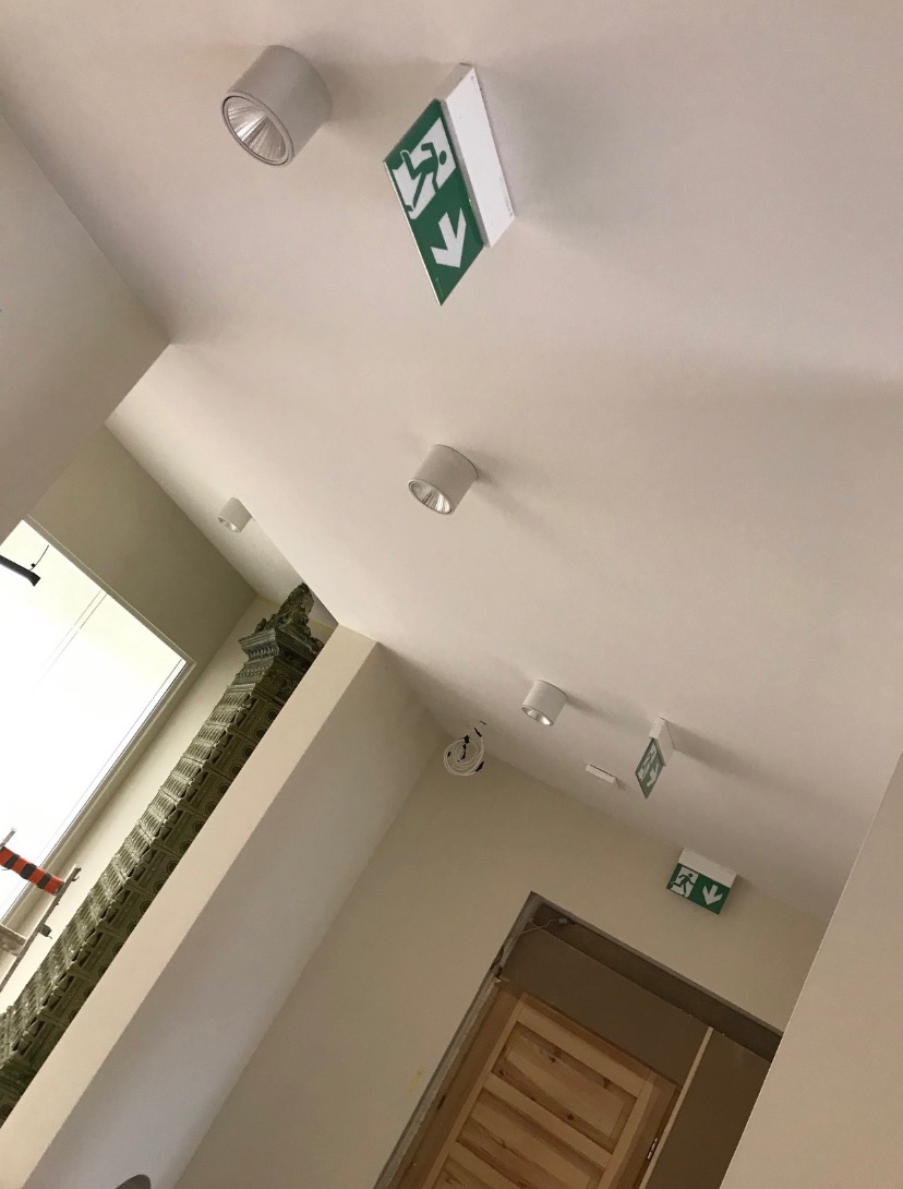 Biały sufit z zamontowanymi okrągłymi lampami i zielonymi znakami ewakuacyjnymi z piktogramem biegnącej osoby i strzałką w dół, w tle fragment ściany z dekoracyjnym elementem.