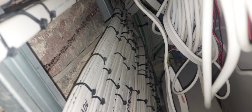 Spięte kable elektryczne w peszlu, prowadzone wzdłuż ściany z widocznymi śladami po instalacji, z dodatkowymi luźnymi przewodami w tle.