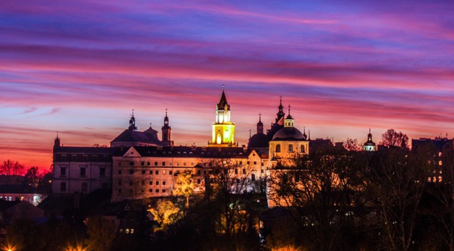 Barokowy kompleks budynków oświetlony nocą, z wyraźnym, wielobarwnym niebem o zachodzie słońca w tle.