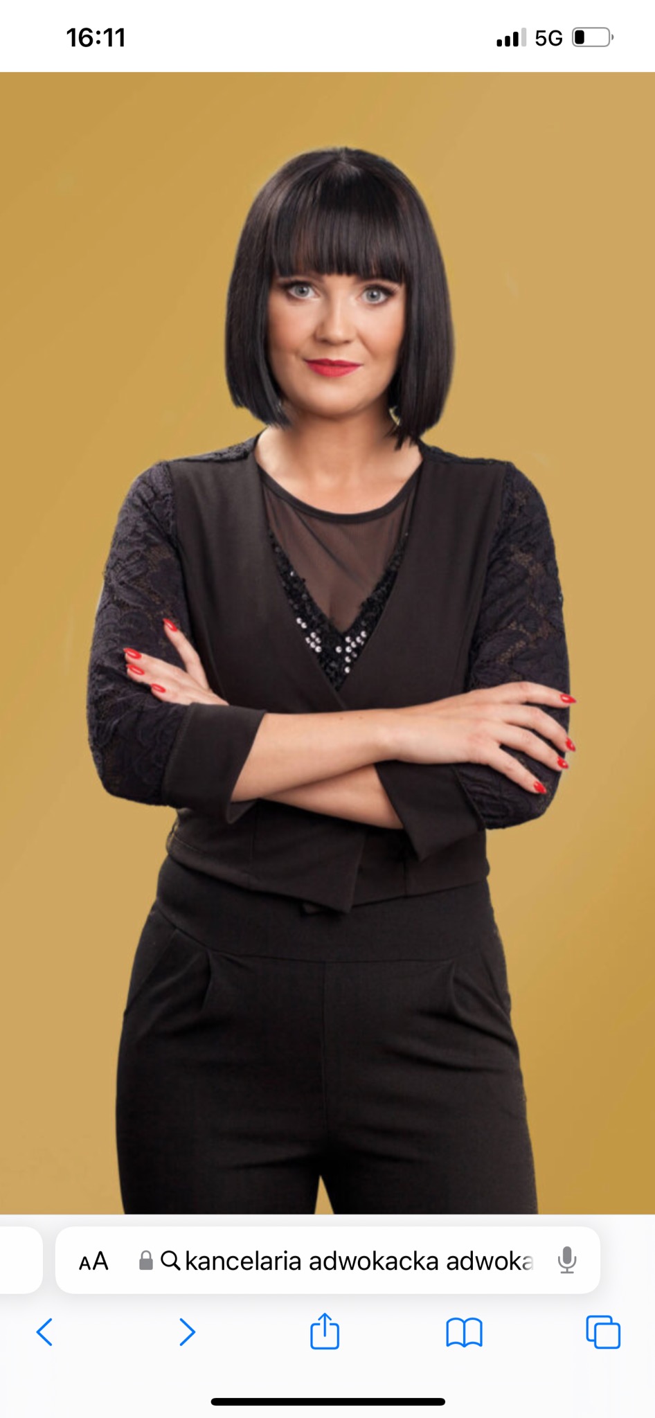 Portret kobiety w czarnym stroju z rękami skrzyżowanymi na piersiach, czerwone paznokcie, bob z grzywką, na jednolitym żółtym tle.