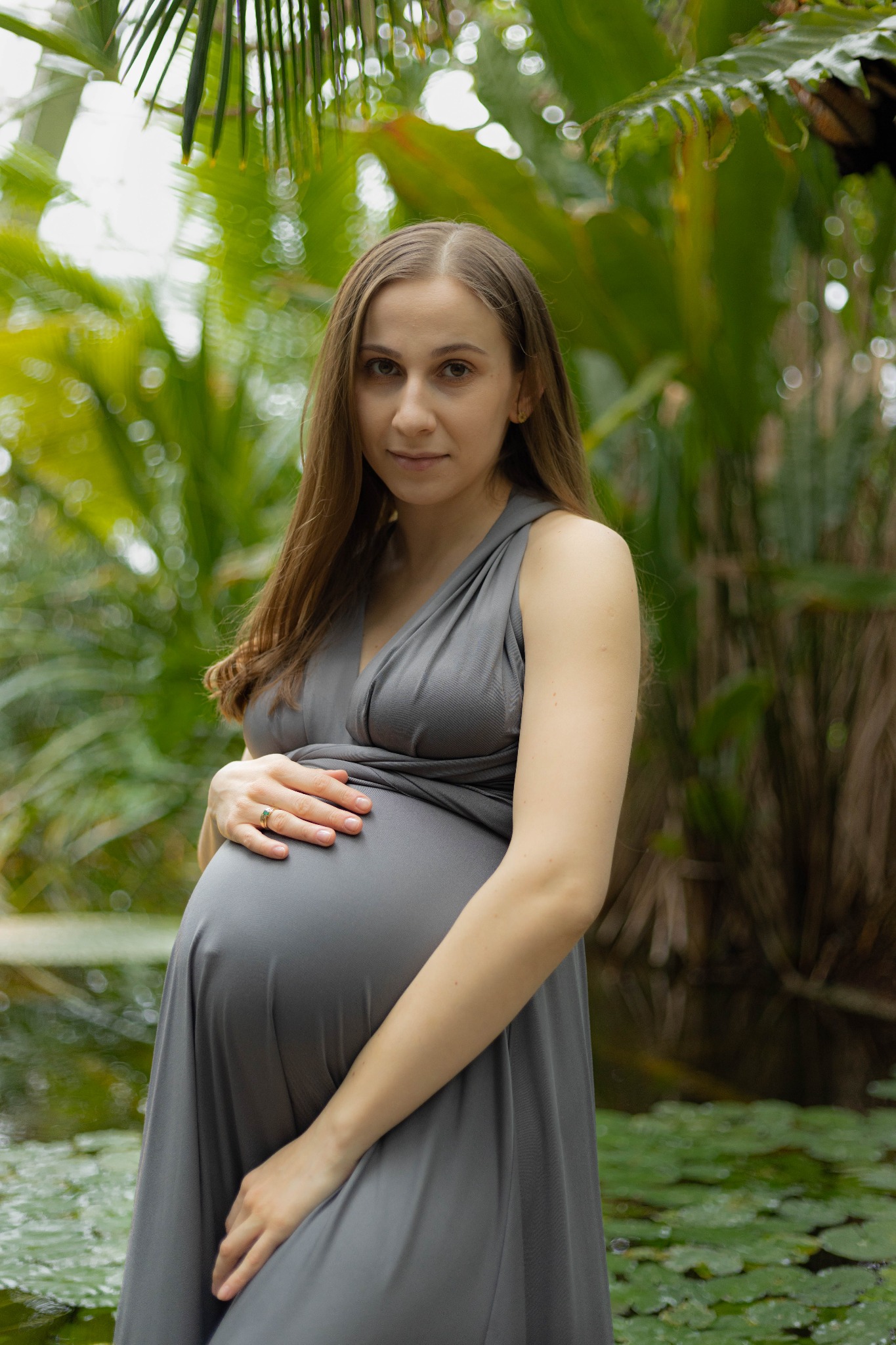Portret kobiety w ciąży w szarej sukni na tle tropikalnej roślinności, dłoń z pierścionkiem na brzuchu.