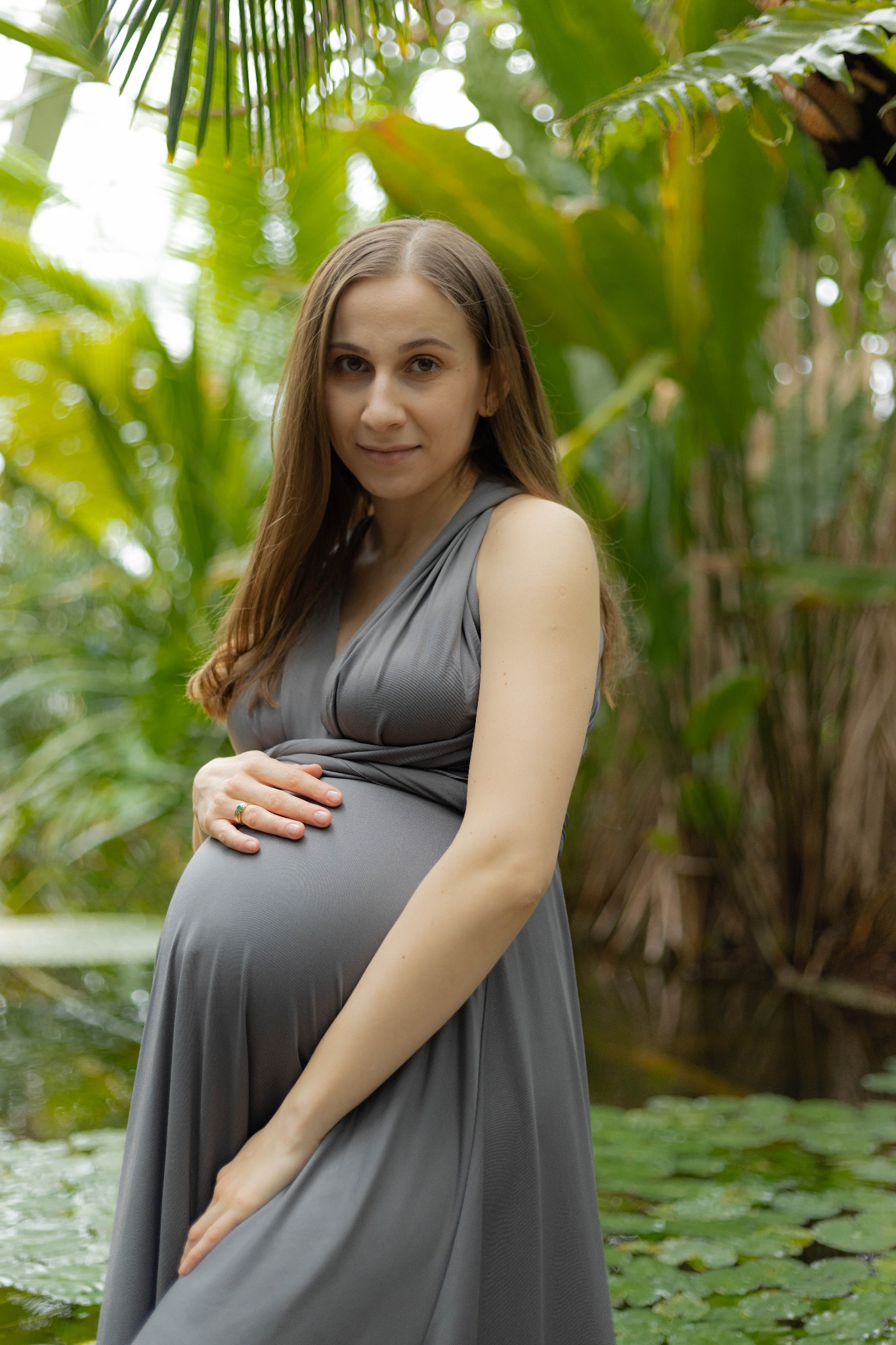 Portret kobiety w ciąży w szarej sukience pozującej na tle zieleni i oczka wodnego, ręce ułożone na brzuchu, pierścionek z zielonym oczkiem na palcu serdecznym.