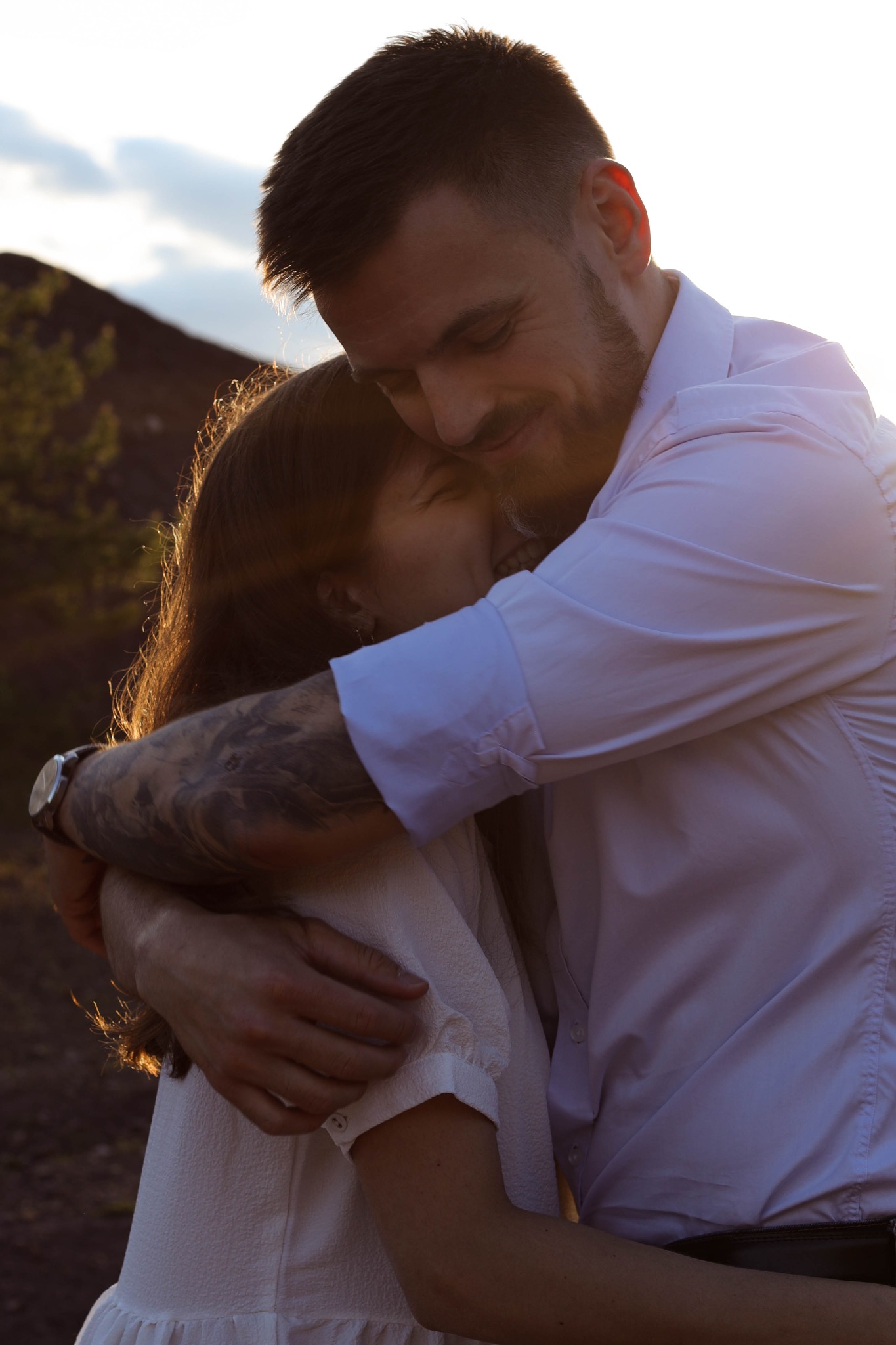 Uśmiechnięty mężczyzna w białej koszuli z tatuażem na ramieniu obejmuje kobietę w jasnej sukience na tle zachodzącego słońca i wzniesień.