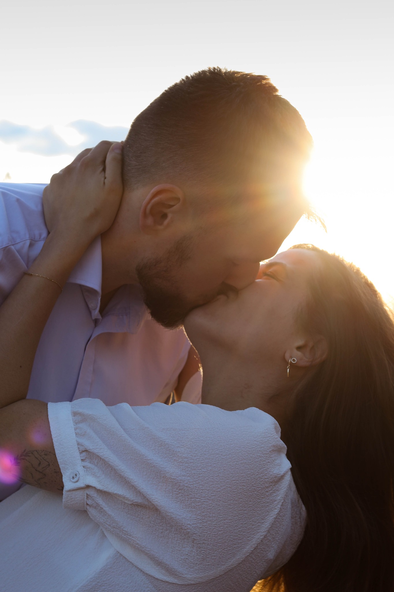 Zakochana para całuje się w silnym, ciepłym świetle słonecznym, widoczny tatuaż na ramieniu kobiety i kolczyk w uchu.