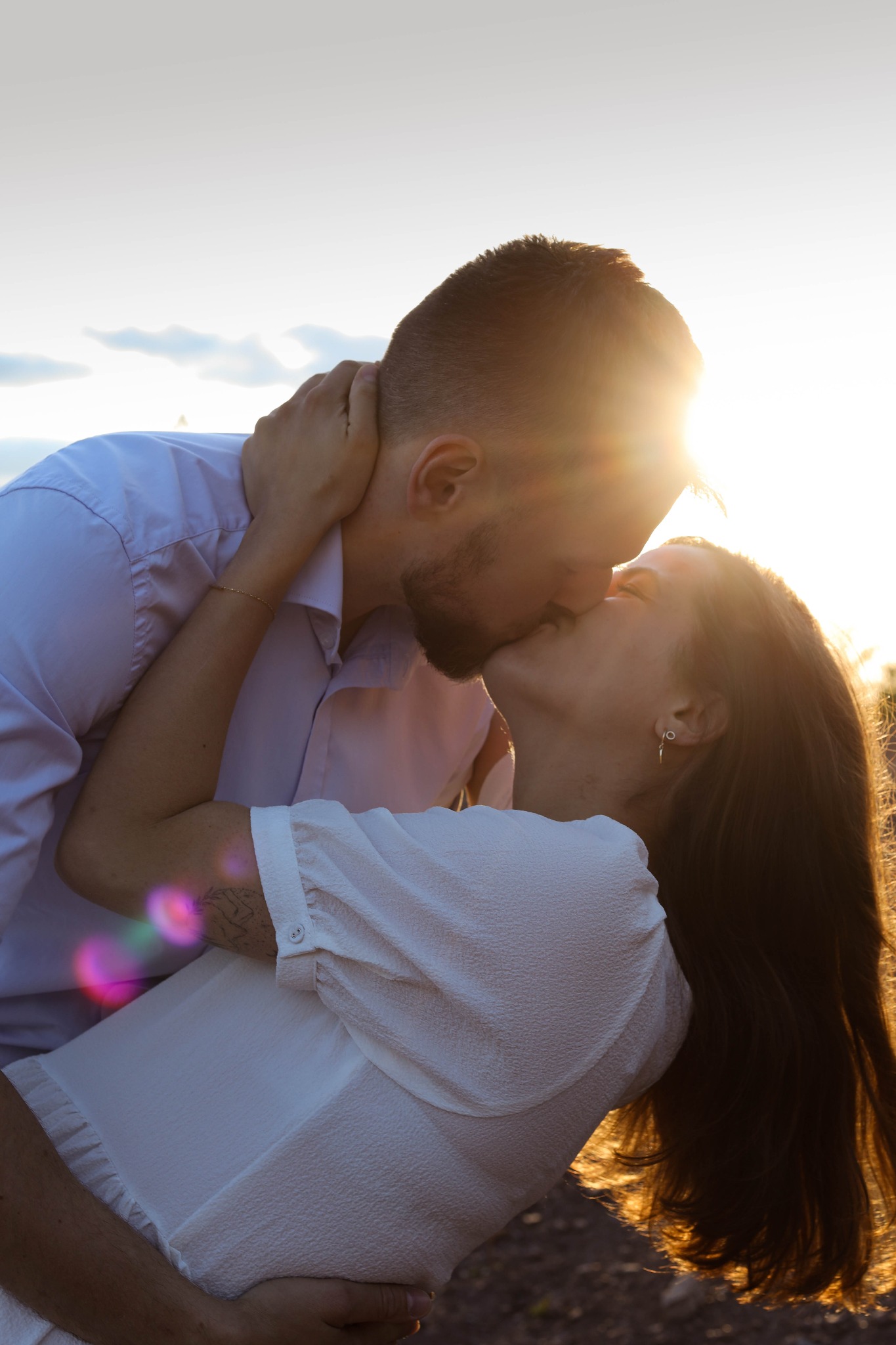 Zakochana para w objęciach, pocałunek w blasku zachodzącego słońca, ona w białej sukience z tatuażem na ramieniu, on w jasnej koszuli.