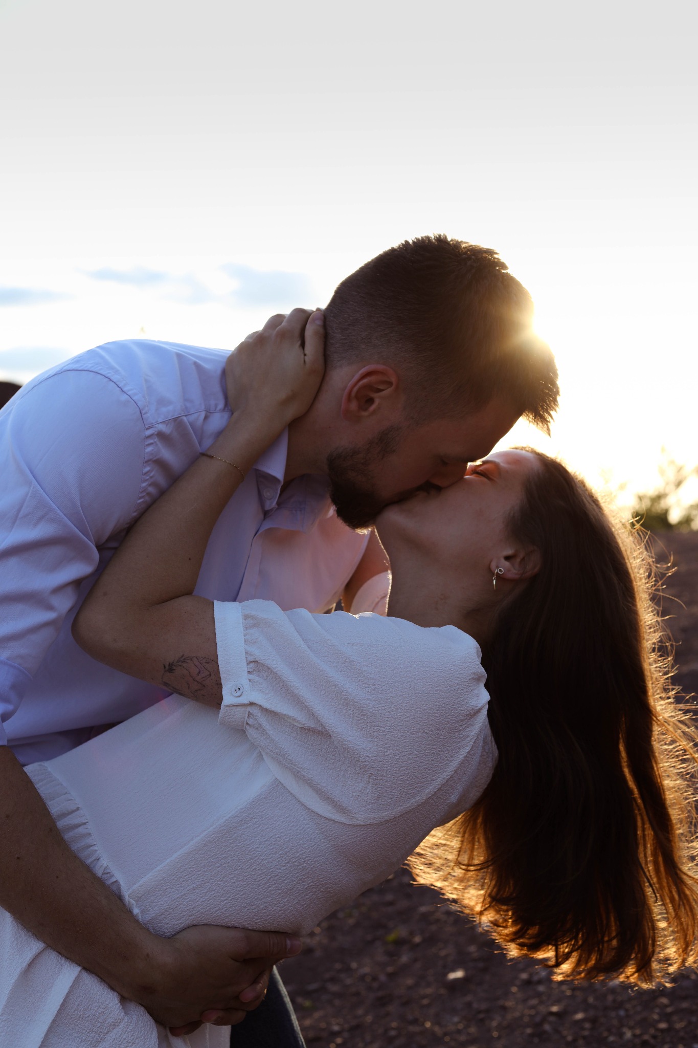 Para całująca się na tle zachodzącego słońca; kobieta w białej sukience z tatuażem na ramieniu, mężczyzna w białej koszuli obejmuje ją. Promienie słońca oświetlają włosy kobiety.