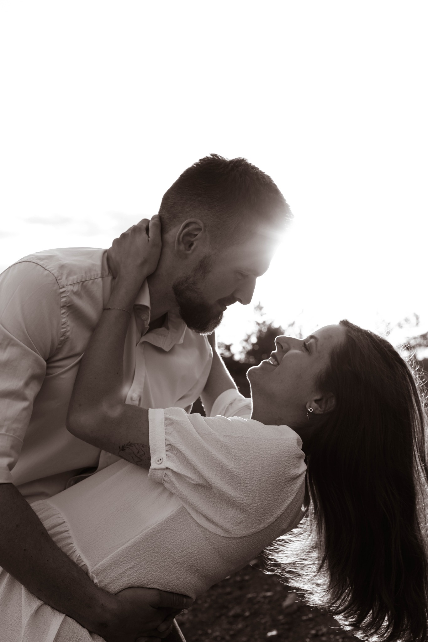 Czarno-białe zdjęcie zakochanej pary, on z brodą obejmuje ją, ona z tatuażem na ramieniu uśmiecha się w jego stronę, w tle rozmyte drzewa i jasne niebo.