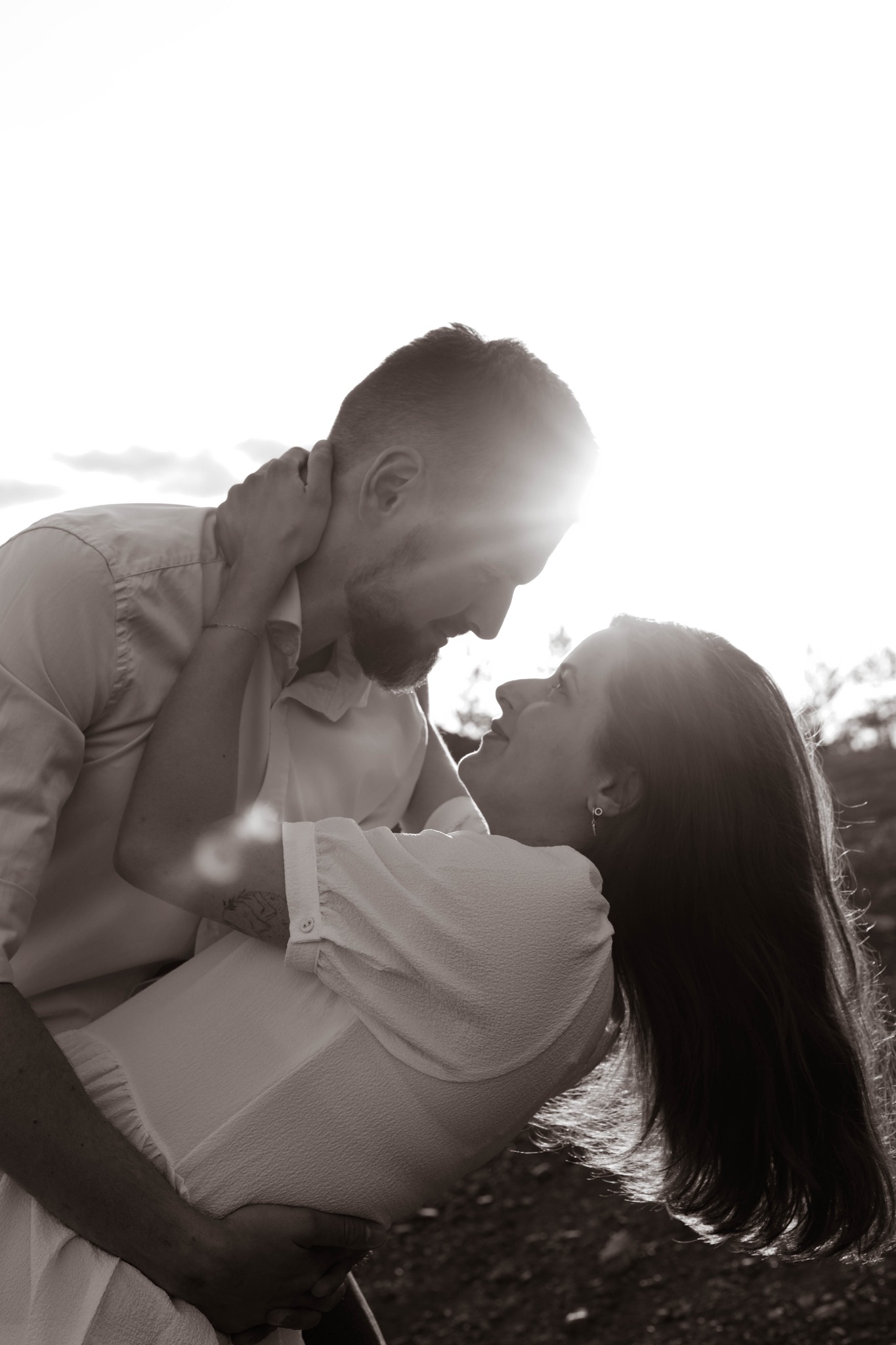 Czarno-białe zdjęcie zakochanej pary, mężczyzna trzyma kobietę w ramionach, oboje patrzą na siebie z uśmiechem, w tle rozproszone światło słoneczne.