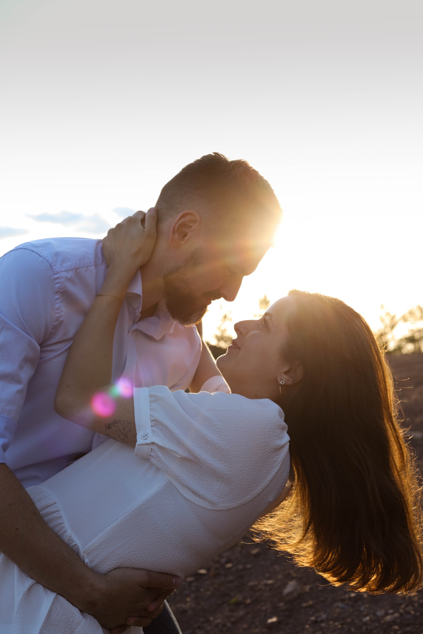 Zakochana para w objęciach, ona w białej sukience, on w koszuli, w kontraście z zachodzącym słońcem i subtelną poświatą na włosach.