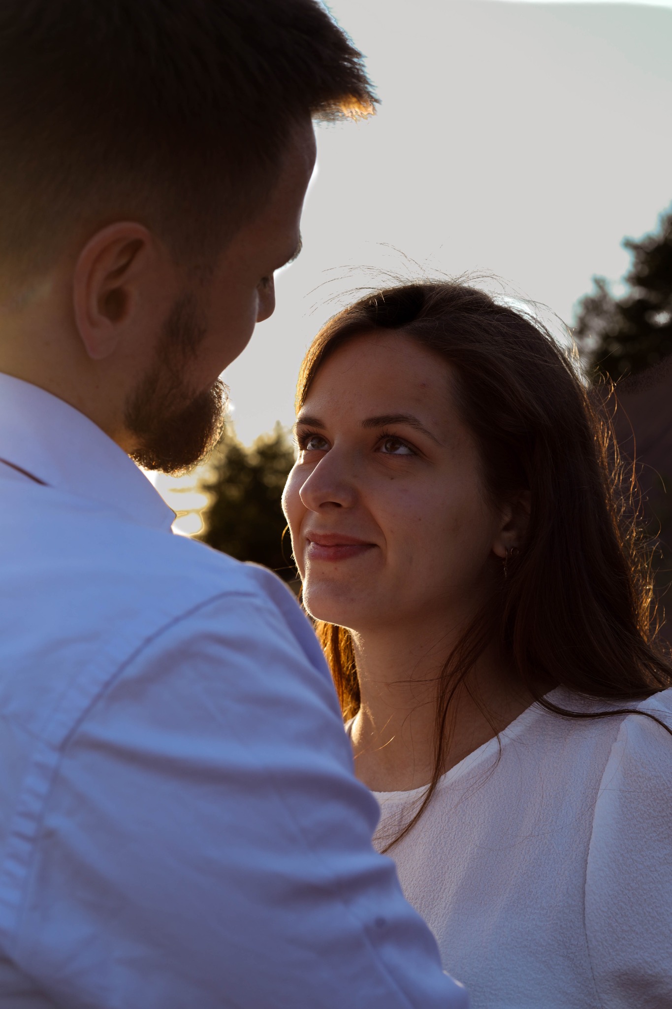 Zakochana para patrzy na siebie podczas sesji narzeczeńskiej w ciepłym świetle zachodzącego słońca, widoczny profil kobiety i fragment brody mężczyzny.