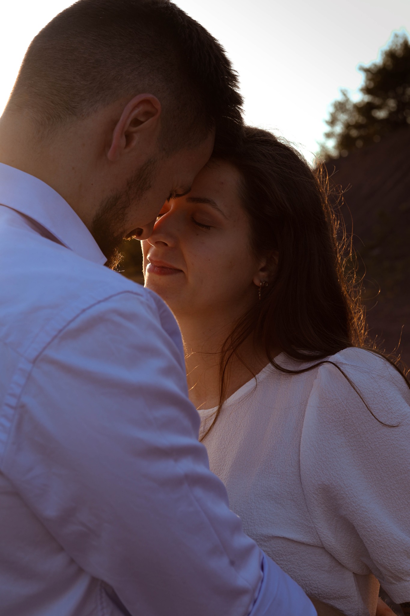 Para zakochanych, zbliżona twarzami, w kontraście z promieniami zachodzącego słońca, kobieta z zamkniętymi oczami i delikatnym uśmiechem.