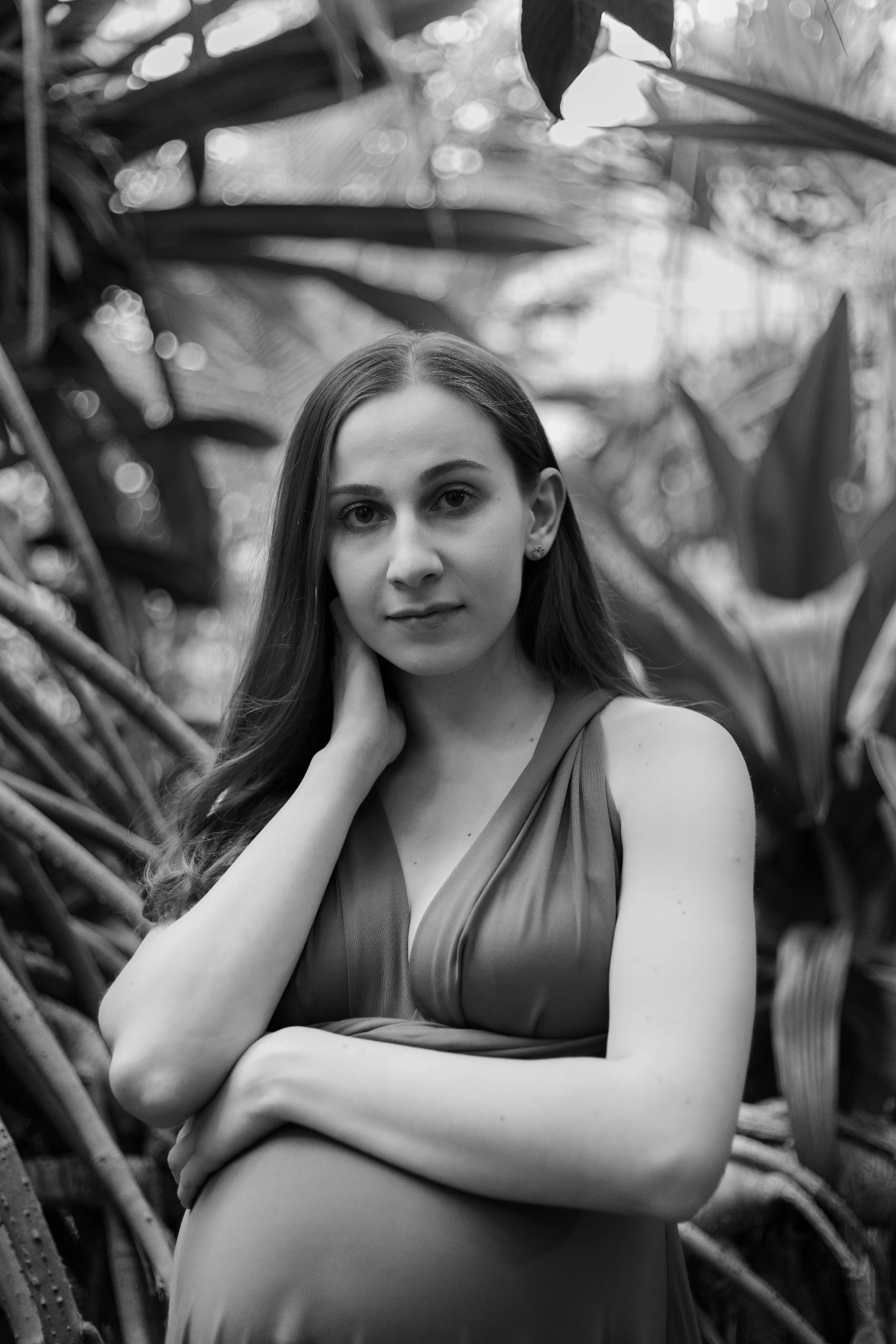 Portret kobiety w sukience, z ręką przy szyi, na tle egzotycznych roślin w czerni i bieli, z delikatnym uśmiechem.