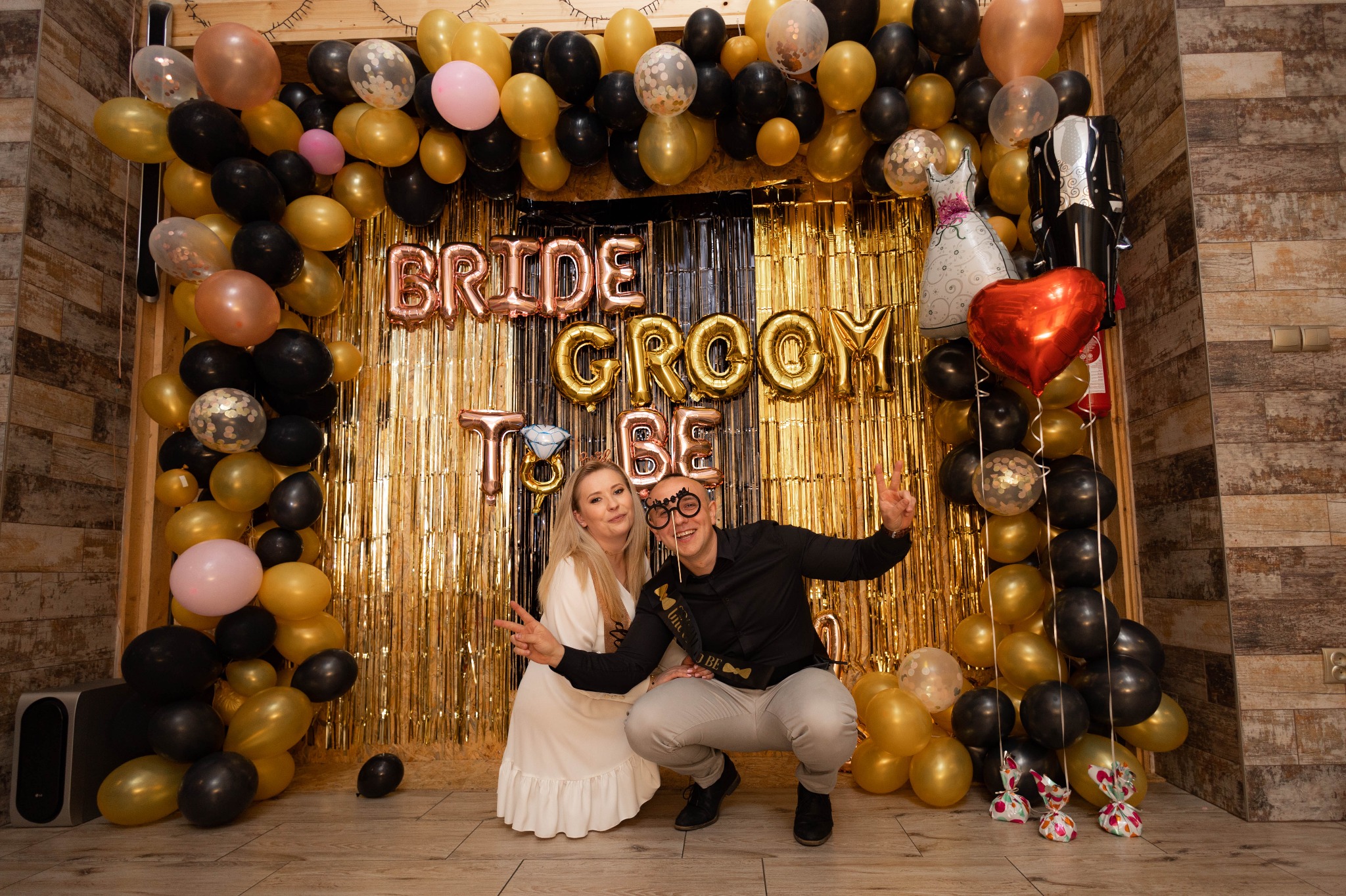 Uśmiechnięta para pozuje na tle dekoracji z balonów i złotych frędzli z napisem Bride to Be, mężczyzna w okularach na patyku i szarfie.