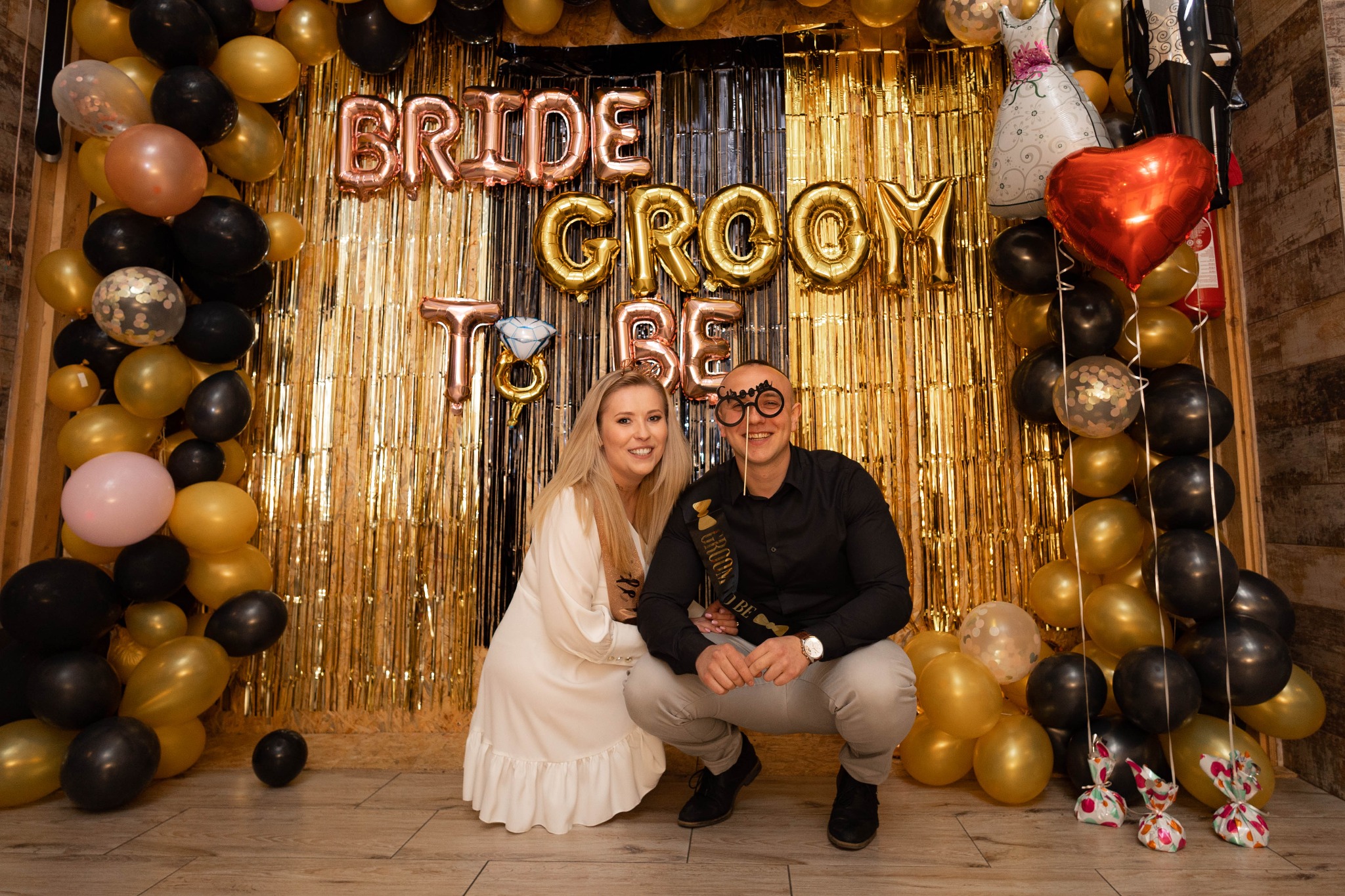 Para narzeczonych w kucającej pozie, z balonami w odcieniach złota i czerni oraz złotą ścianką dekoracyjną z napisem 'Bride Groom To Be', mężczyzna z rekwizytami na patyku w kształcie okularów...