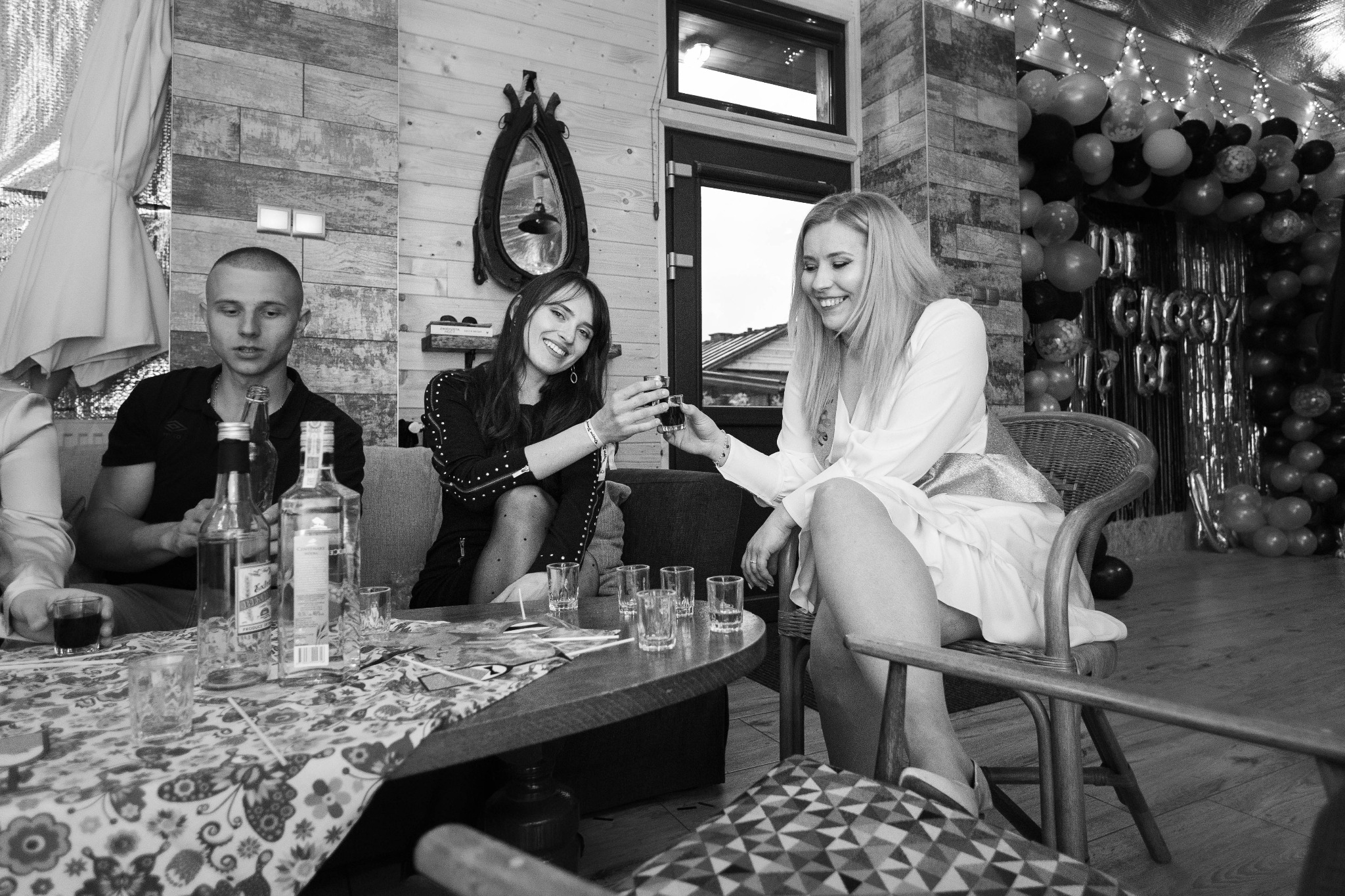 Czarno-białe zdjęcie przedstawia troje ludzi siedzących przy stole z kieliszkami i butelkami, w tle drewniana ściana, lustro i dekoracje balonowe, sugerujące imprezę.