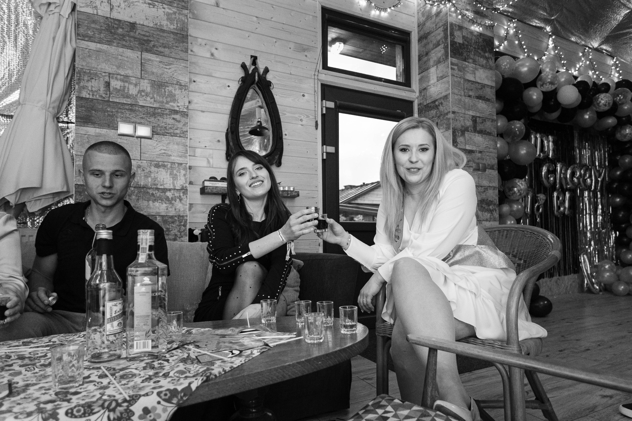 Czarno-białe zdjęcie trzech osób w pomieszczeniu udekorowanym balonami, siedzących przy stole z butelkami i kieliszkami, wznoszących toast. W tle drewniane ściany i dekoracyjne oświetlenie.